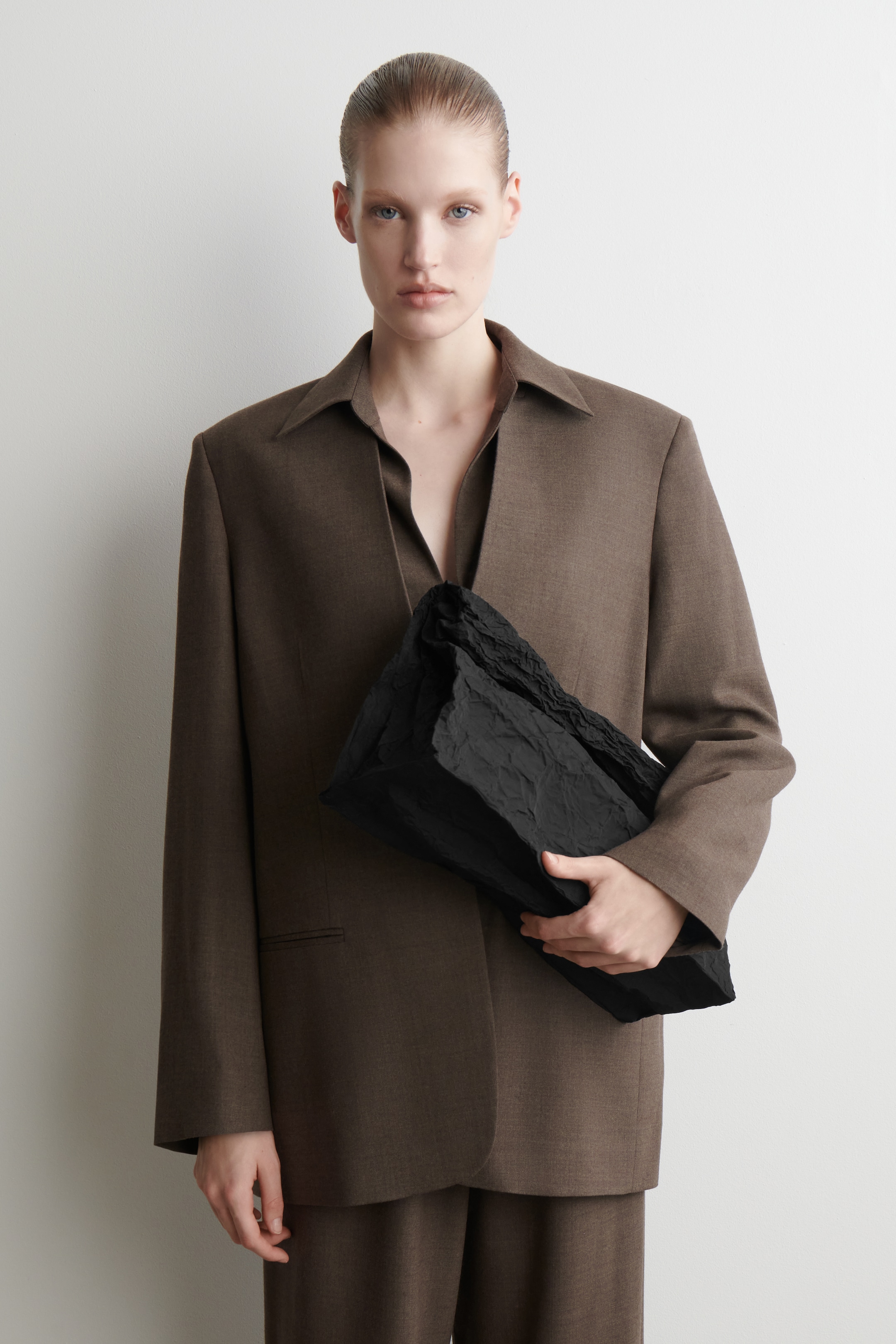 Visualizza immagine più grande: Il blazer marrone viene indossato, mettendo in mostra la sua vestibilità rilassata e oversize con spalle larghe e un ampio colletto aperto. Presenta un'apertura frontale e una singola tasca a patta, abbinato a un top marrone a V e pantaloni coordinati, con una pochette nera stropicciata tenuta contro di esso.