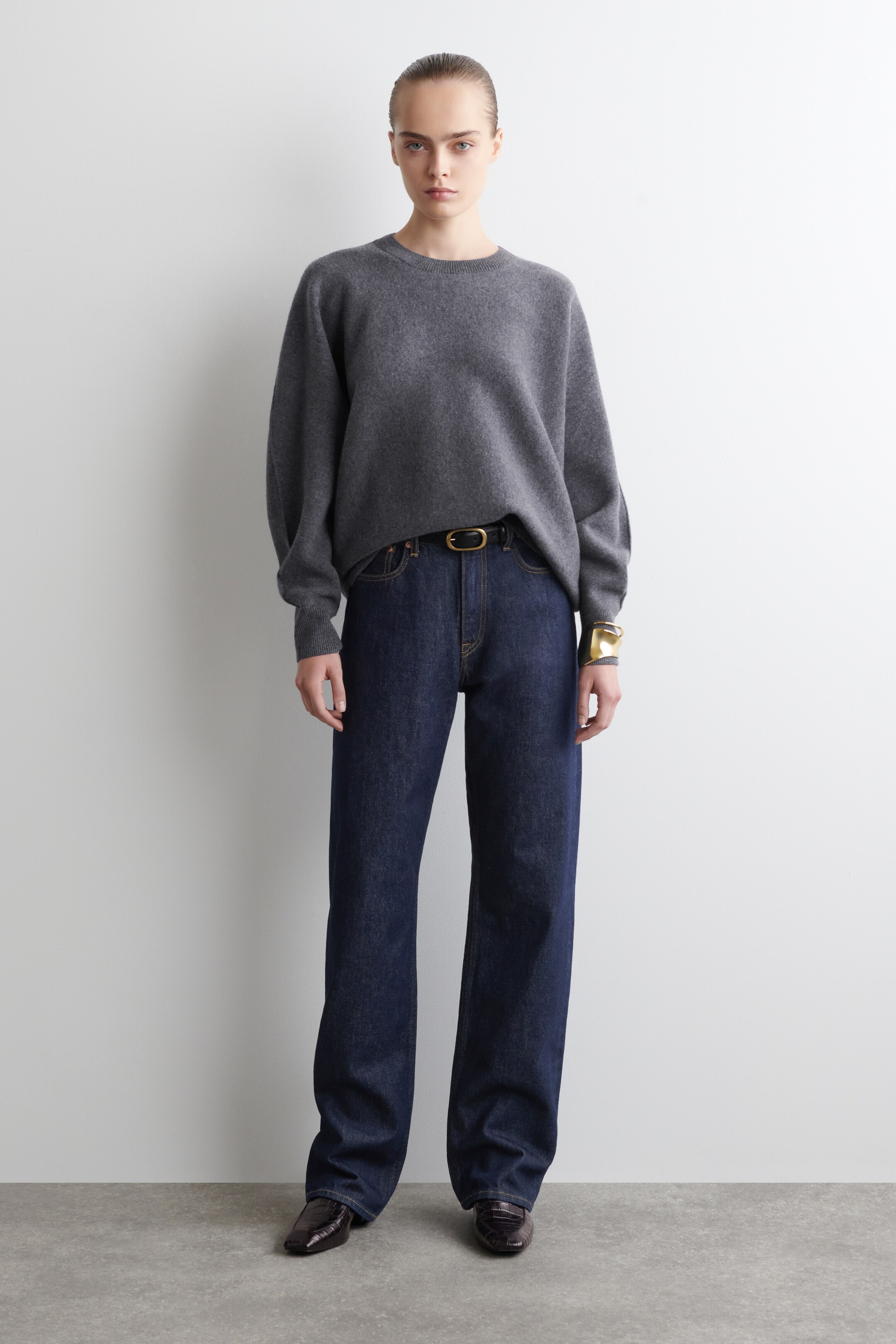 Ingrandisci l'immagine: Un maglione grigio scuro a girocollo con una vestibilità rilassata e maniche voluminose che terminano in polsini a coste è indossato. È abbinato a jeans a gamba larga con lavaggio scuro, caratterizzati da cuciture gialle prominenti, una cintura nera con fibbia dorata e mocassini marrone scuro a punta, con un braccialetto rigido dorato visibile anche al polso.