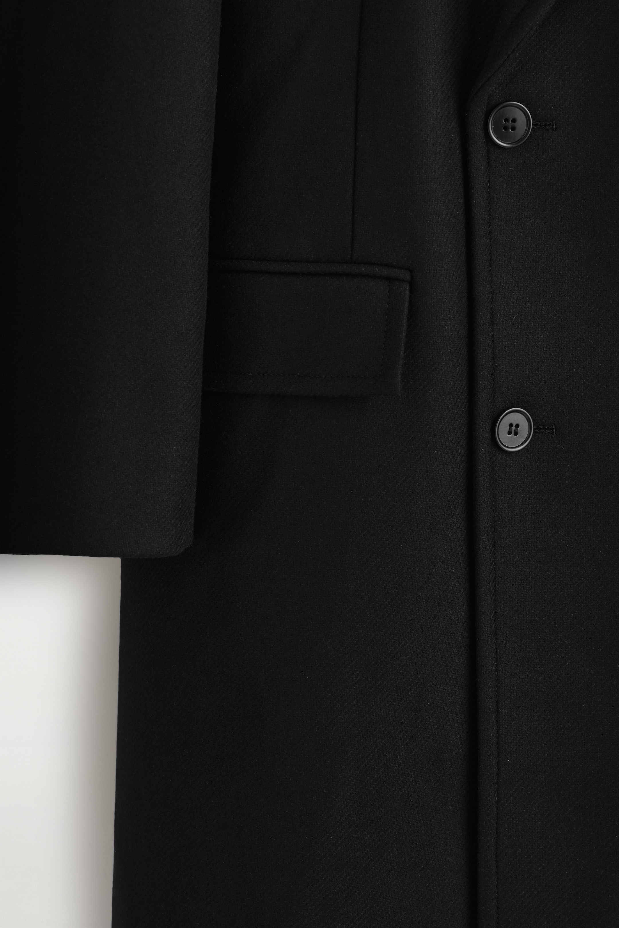 Visualizza immagine più grande: La parte anteriore di un cappotto nero è mostrata in primo piano, con due bottoni neri a quattro fori con asole e una tasca a patta rettangolare. Il tessuto presenta una sottile trama diagonale.