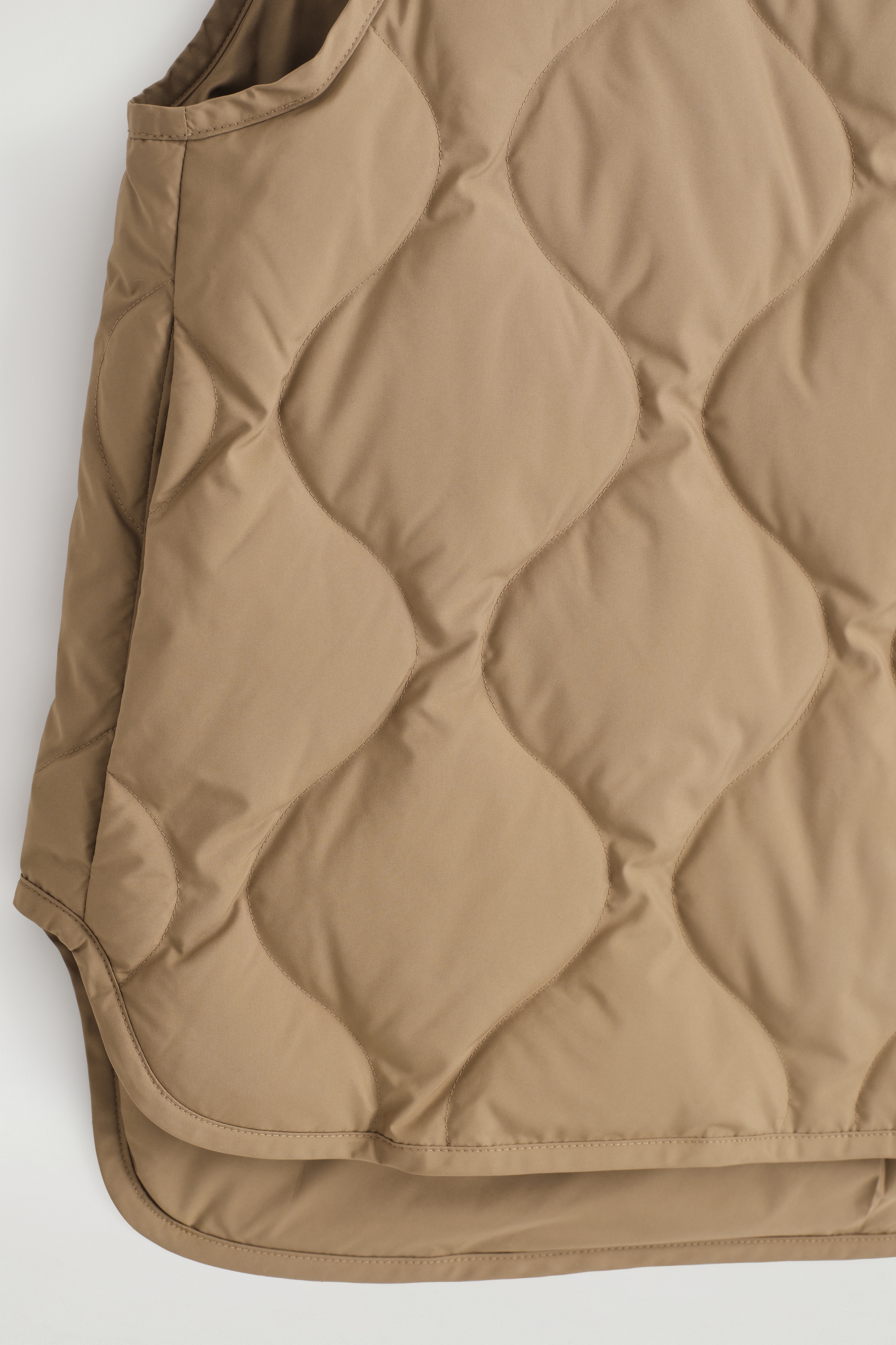 View larger image: The front body of a caramel-colored gilet is displaying a padded texture with large, wavy quilted patterns. An armhole opening is visible on the left, and the gilet is featuring a rounded hemline at the bottom.