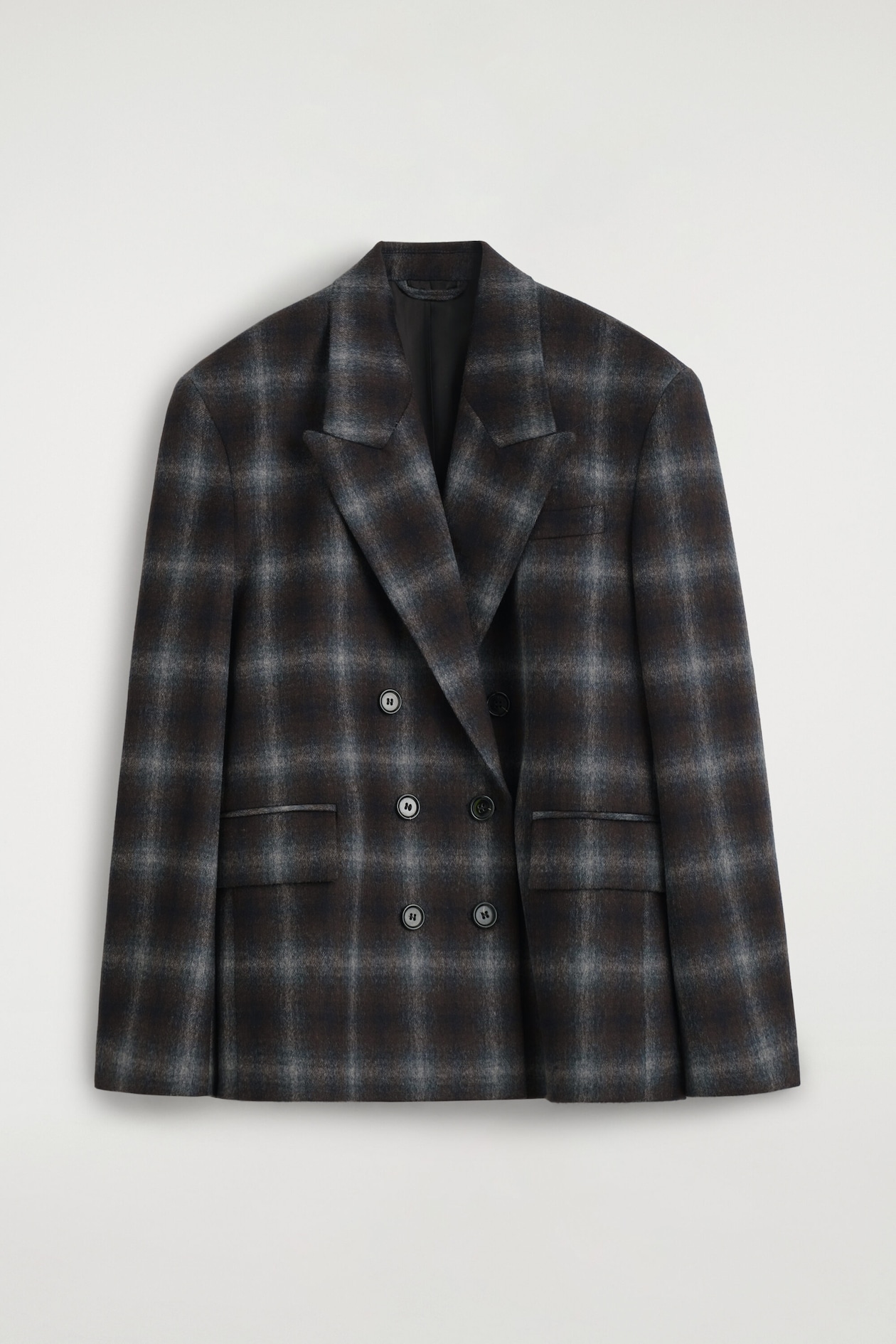 Ce blazer croisé en mélange de laine à carreaux affiche un motif à carreaux marron foncé et gris vu de face. Le blazer présente de larges revers à pointes, une fermeture croisée avec des boutons foncés et deux poches à rabat à l’avant.