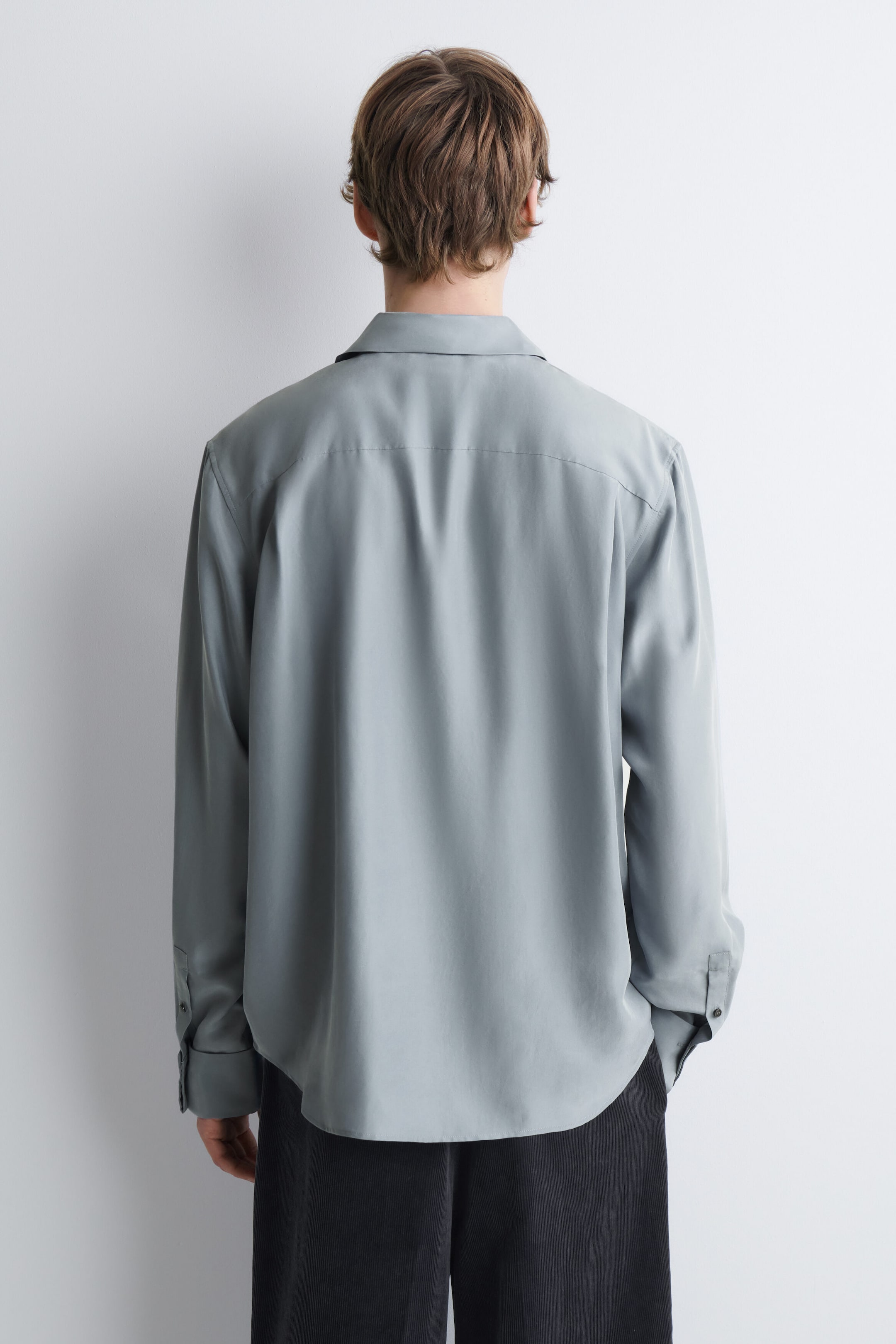 A relaxed silk shirt in a pale grey-green hue is worn by the model, shown from the back. The shirt features a classic collar, a straight back yoke, long sleeves with buttoned cuffs, and a slightly curved hem. A relaxed silk shirt in a pale grey-green hue is worn by the model, shown from the back. The shirt features a classic collar, a straight back yoke, long sleeves with buttoned cuffs, and a slightly curved hem.
