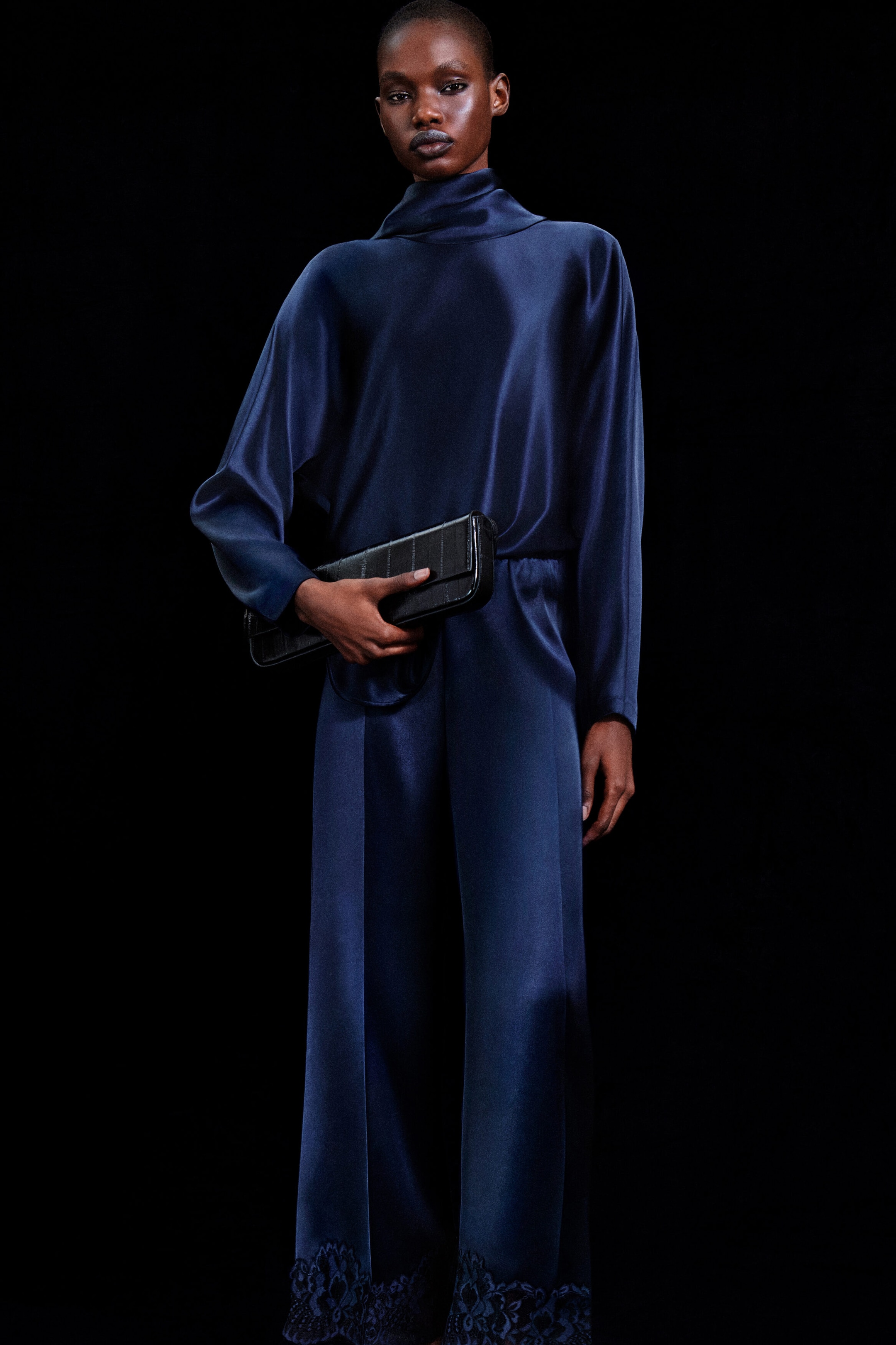 A model facing the camera is wearing a navy blue cowl-neck satin blouse featuring a high draped collar and loose long sleeves. The blouse is paired with matching wide-leg satin trousers showcasing intricate lace trim along the bottom hems. A model facing the camera is wearing a navy blue cowl-neck satin blouse featuring a high draped collar and loose long sleeves. The blouse is paired with matching wide-leg satin trousers showcasing intricate lace trim along the bottom hems.