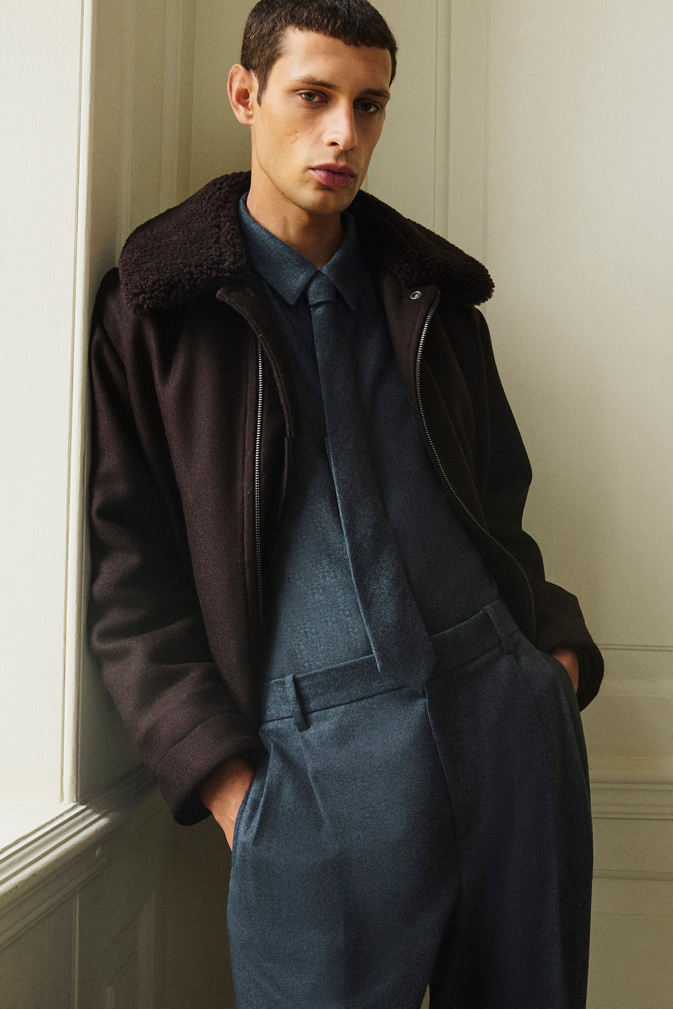 The relaxed wool flannel shirt in blue-grey is shown from the front, styled with a matching tie and tucked into coordinating trousers. It is layered beneath an open dark brown flight jacket with a shearling collar. The relaxed wool flannel shirt in blue-grey is shown from the front, styled with a matching tie and tucked into coordinating trousers. It is layered beneath an open dark brown flight jacket with a shearling collar.