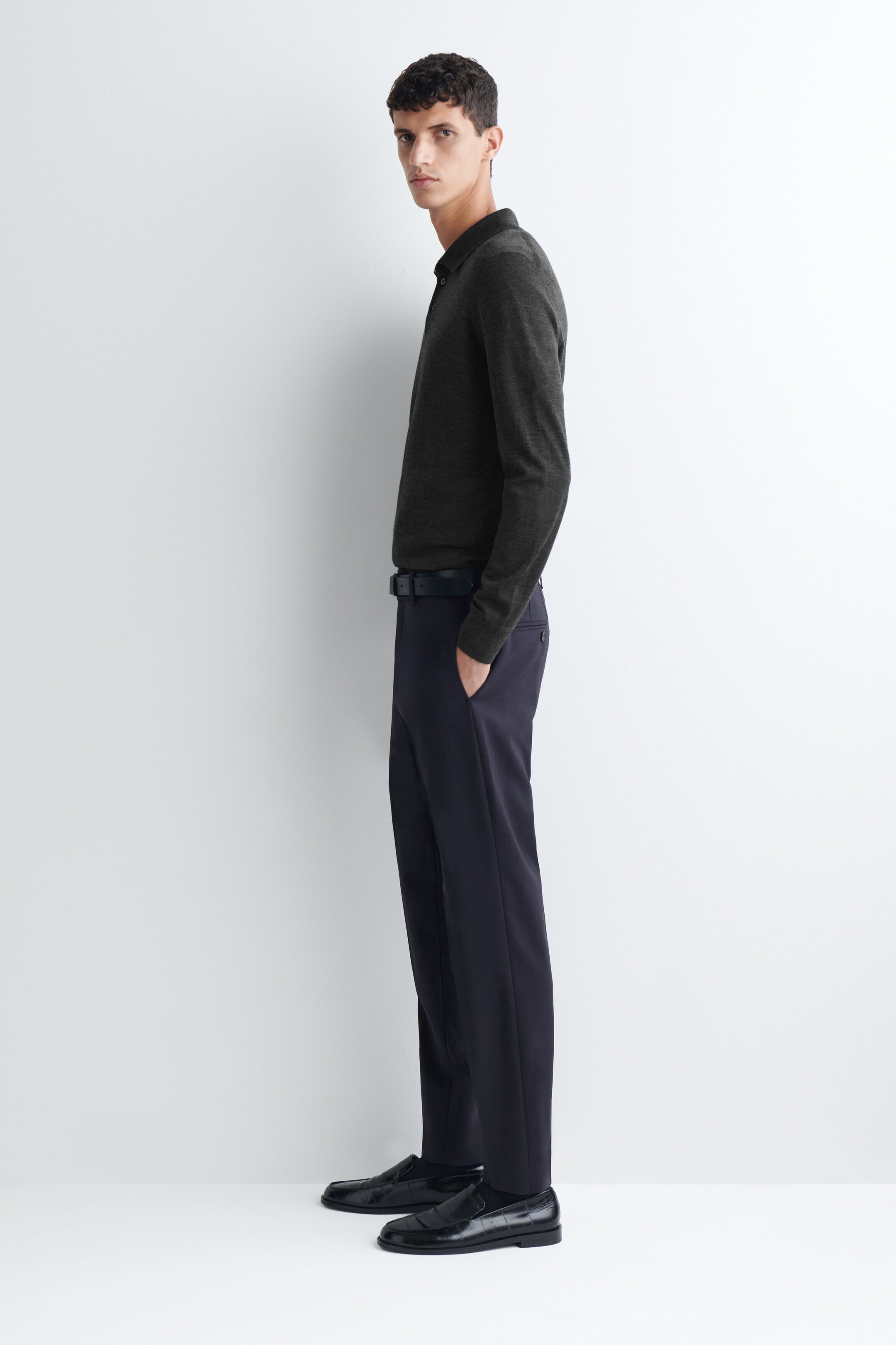 Le mannequin porte un pantalon droit slim en laine bleu marine foncé, en position de profil, avec une main glissée dans la poche latérale. Le pantalon présente une ceinture structurée maintenue par une ceinture noire, un pli net marqué le long de la jambe, et est associé à un polo à manches longues gris foncé rentré à l’intérieur ainsi qu’à des mocassins en cuir noir. Le mannequin porte un pantalon droit slim en laine bleu marine foncé, en position de profil, avec une main glissée dans la poche latérale. Le pantalon présente une ceinture structurée maintenue par une ceinture noire, un pli net marqué le long de la jambe, et est associé à un polo à manches longues gris foncé rentré à l’intérieur ainsi qu’à des mocassins en cuir noir.