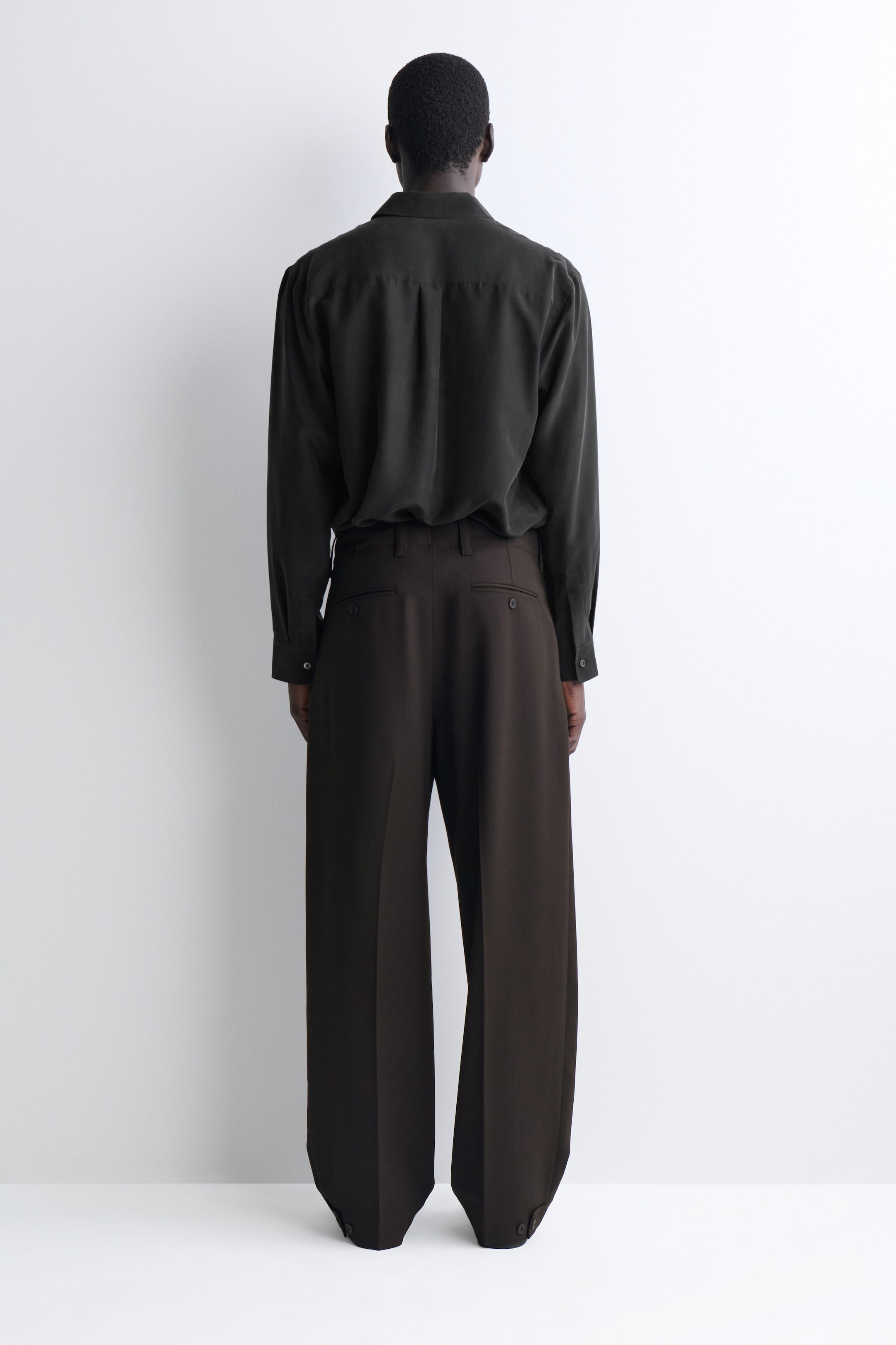 A dark olive-green long-sleeve shirt and dark brown wide-leg trousers are shown from the back to highlight their relaxed fit and design. The draped shirt is tucked into the trousers, which feature buttoned back welt pockets, centre leg creases, and adjustable buttoned tabs at the hems. A dark olive-green long-sleeve shirt and dark brown wide-leg trousers are shown from the back to highlight their relaxed fit and design. The draped shirt is tucked into the trousers, which feature buttoned back welt pockets, centre leg creases, and adjustable buttoned tabs at the hems.