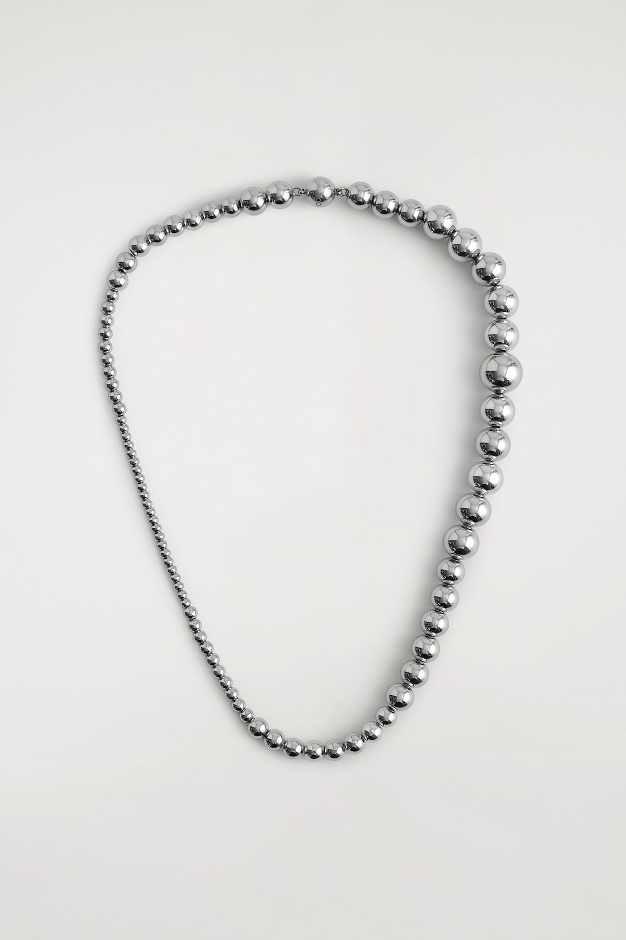 Un collier de perles est posé à plat, mettant en valeur des perles sphériques à finition argentée polie de tailles dégradées, disposées en boucle asymétrique. Les perles passent de très petites sphères d’un côté à des sphères plus grandes, hautement réfléchissantes, de l’autre.
