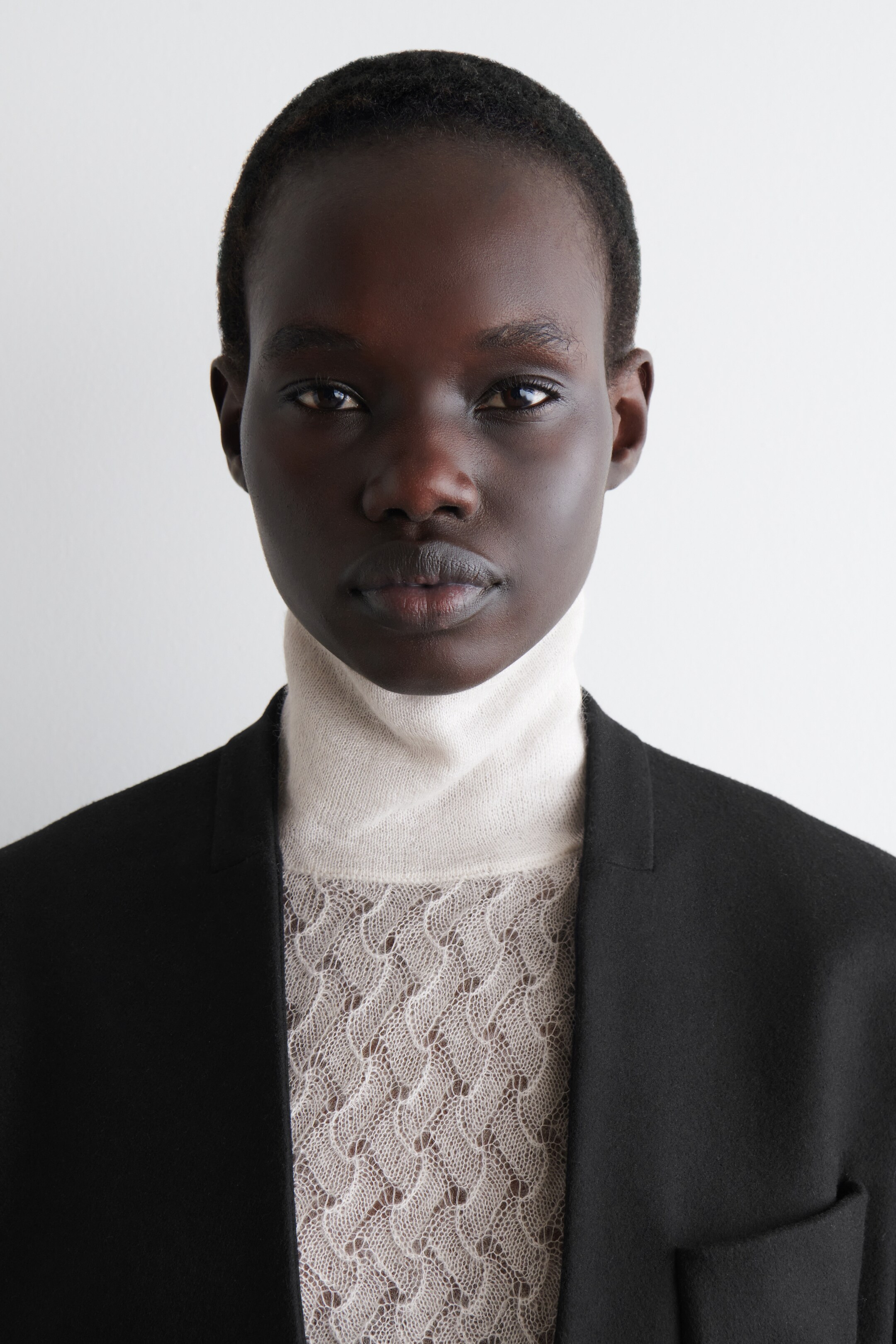 Visa större bild: En ljusbeige långärmad TOP har en ribbad trattkrage. Dess kropp visar ett delikat pointelle-stickmönster och bärs lager-på-lager under en svart blazer.