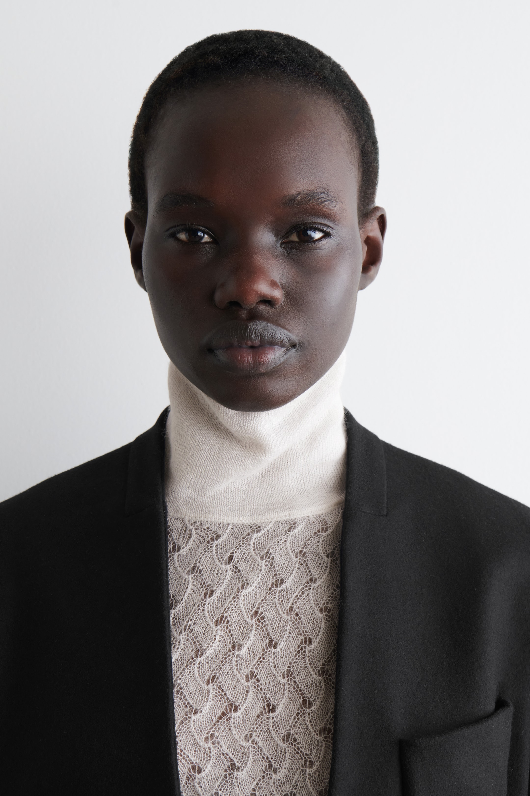 Grösseres Bild ansehen: Ein hellbeiges langärmliges TOP mit einem gerippten Trichterkragen. Der Körper zeigt ein zartes Pointelle-Strickmuster und wird unter einem schwarzen Blazer getragen.
