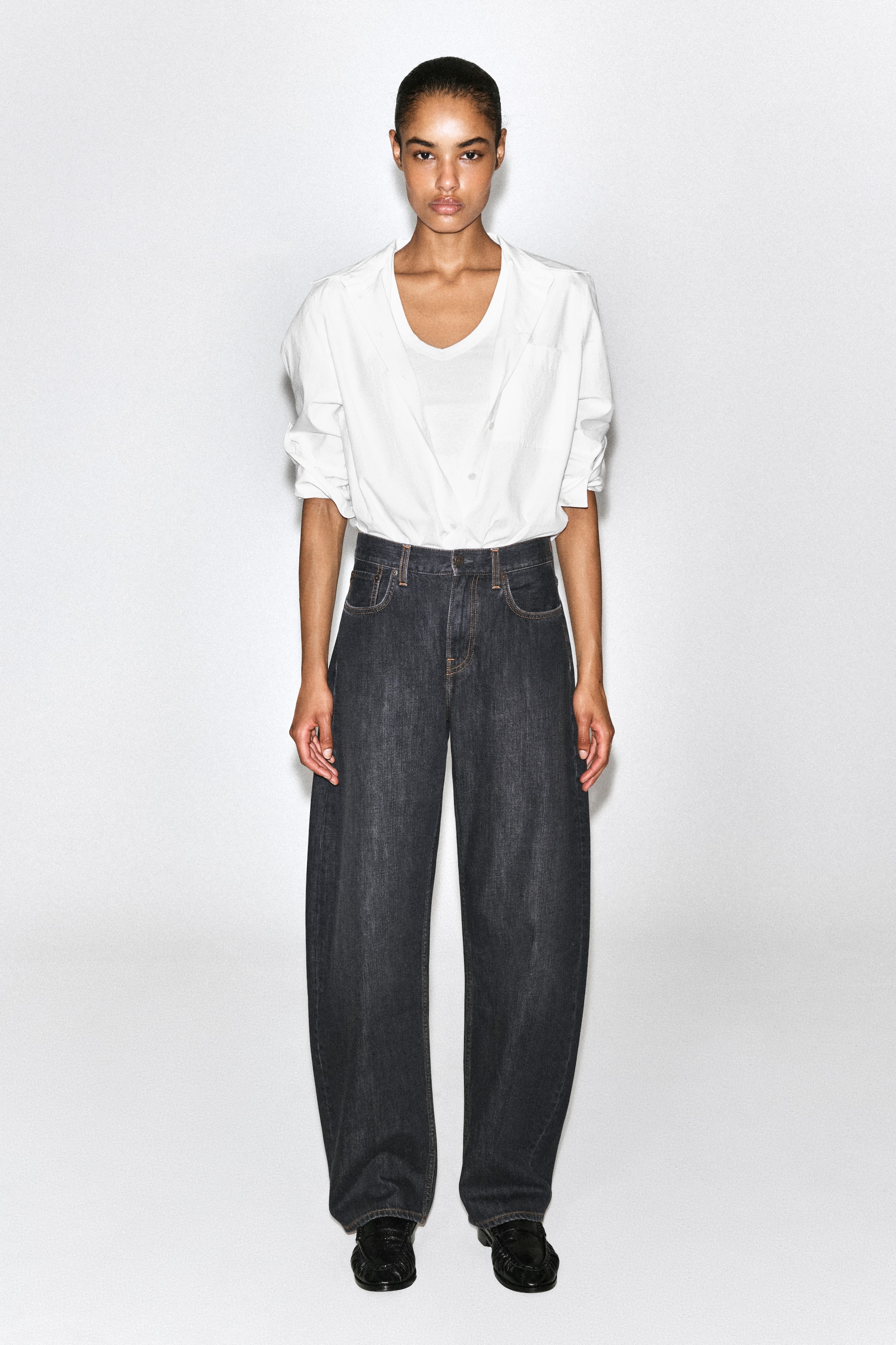 Ver imagen más grande: Los vaqueros de lavado oscuro y pernera curvada se presentan de frente sobre una modelo, mostrando su corte relajado, tiro medio y el clásico diseño de cinco bolsillos con pespuntes en contraste. Una camisa blanca metida por dentro deja ver la cinturilla, resaltando la distintiva silueta arquitectónica de los vaqueros.