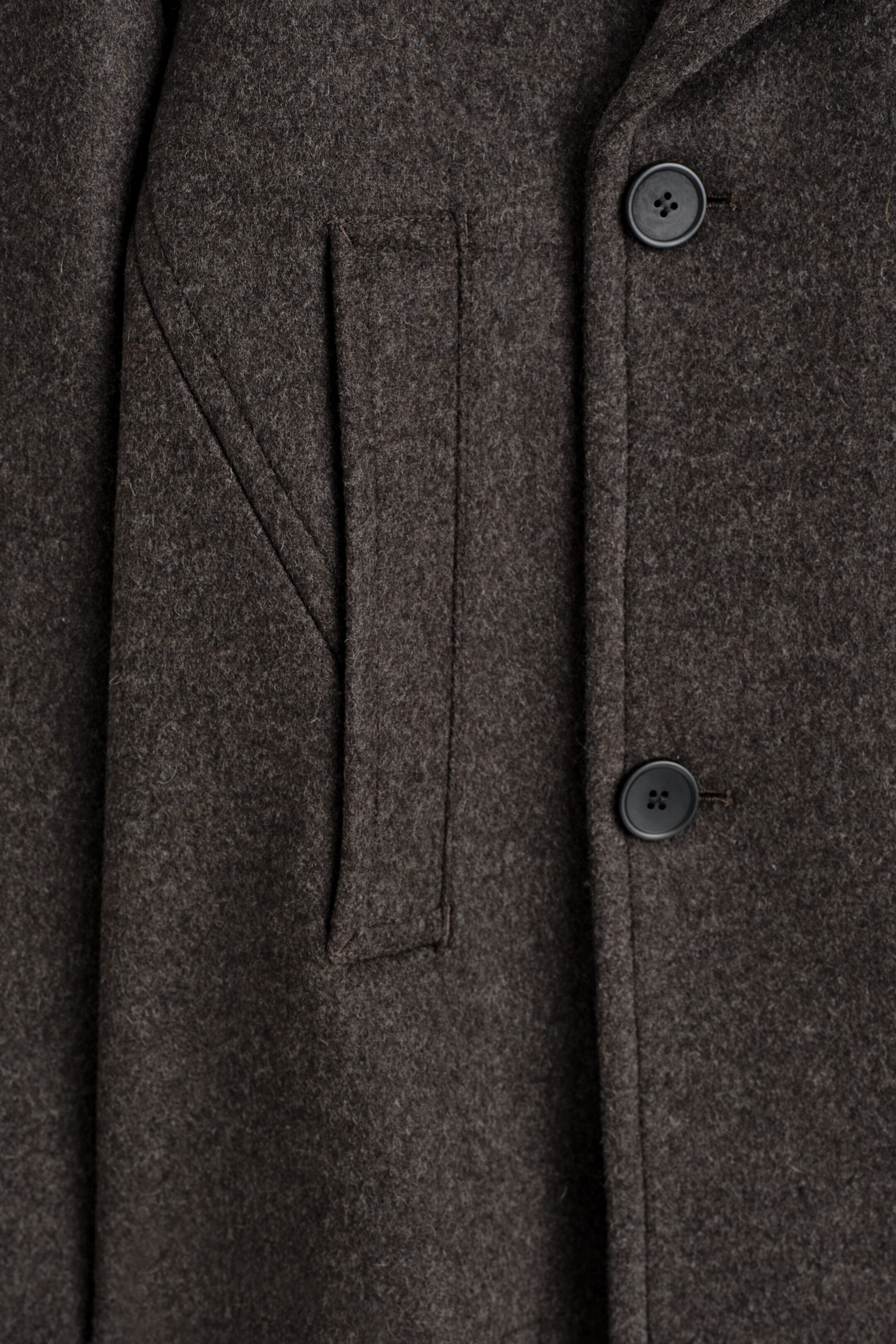 Visualizza immagine più grande: La PARTE ANTERIORE di un cappotto, realizzato in lana tattile di un ricco tono espresso, presenta un bottone nero e una tasca a filetto larga con cuciture distintive.