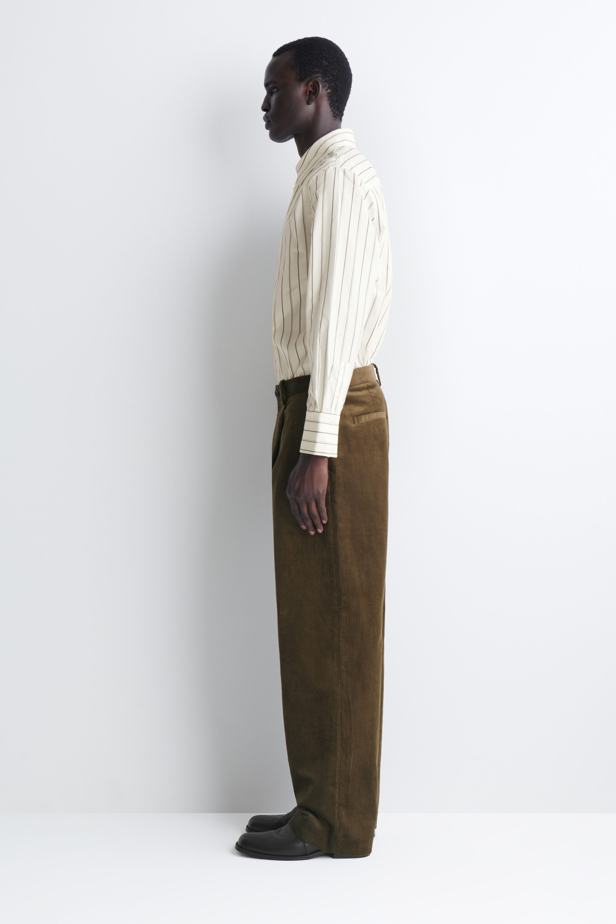 Pleated relaxed-fit tapered corduroy pants in a textured olive-brown fabric are shown in profile facing left, highlighting their relaxed silhouette and slight taper. The pants feature a structured waistband with a tucked-in striped shirt and drape over black leather shoes. Pleated relaxed-fit tapered corduroy pants in a textured olive-brown fabric are shown in profile facing left, highlighting their relaxed silhouette and slight taper. The pants feature a structured waistband with a tucked-in striped shirt and drape over black leather shoes.