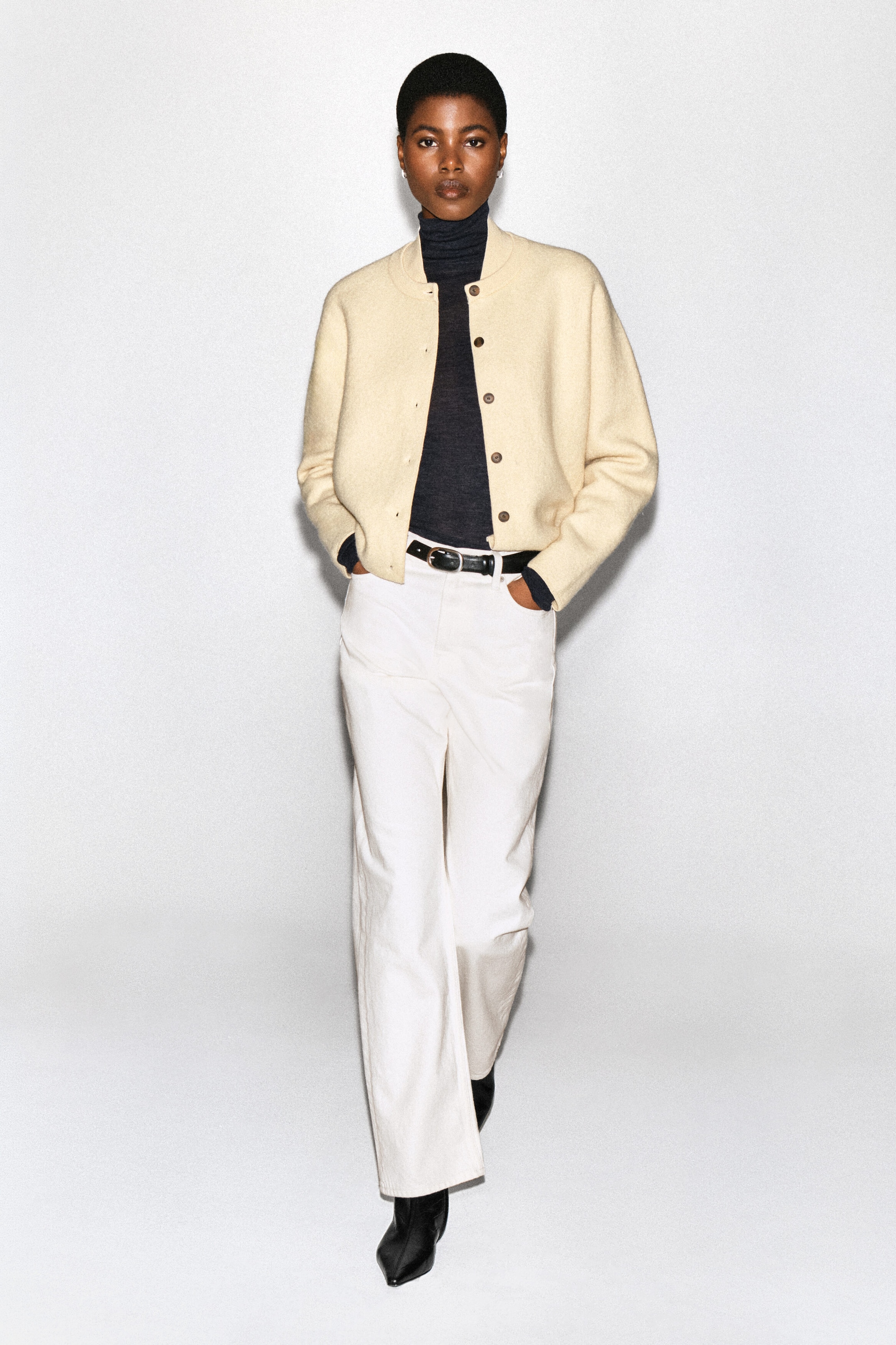 Un cardigan en laine bouillie couleur crème est porté par le mannequin, superposé sur un haut col roulé gris foncé. Le cardigan présente une silhouette arrondie, un col de style bomber et des boutons foncés à l’avant, et il est agencé avec un pantalon blanc et une ceinture noire. Un cardigan en laine bouillie couleur crème est porté par le mannequin, superposé sur un haut col roulé gris foncé. Le cardigan présente une silhouette arrondie, un col de style bomber et des boutons foncés à l’avant, et il est agencé avec un pantalon blanc et une ceinture noire.