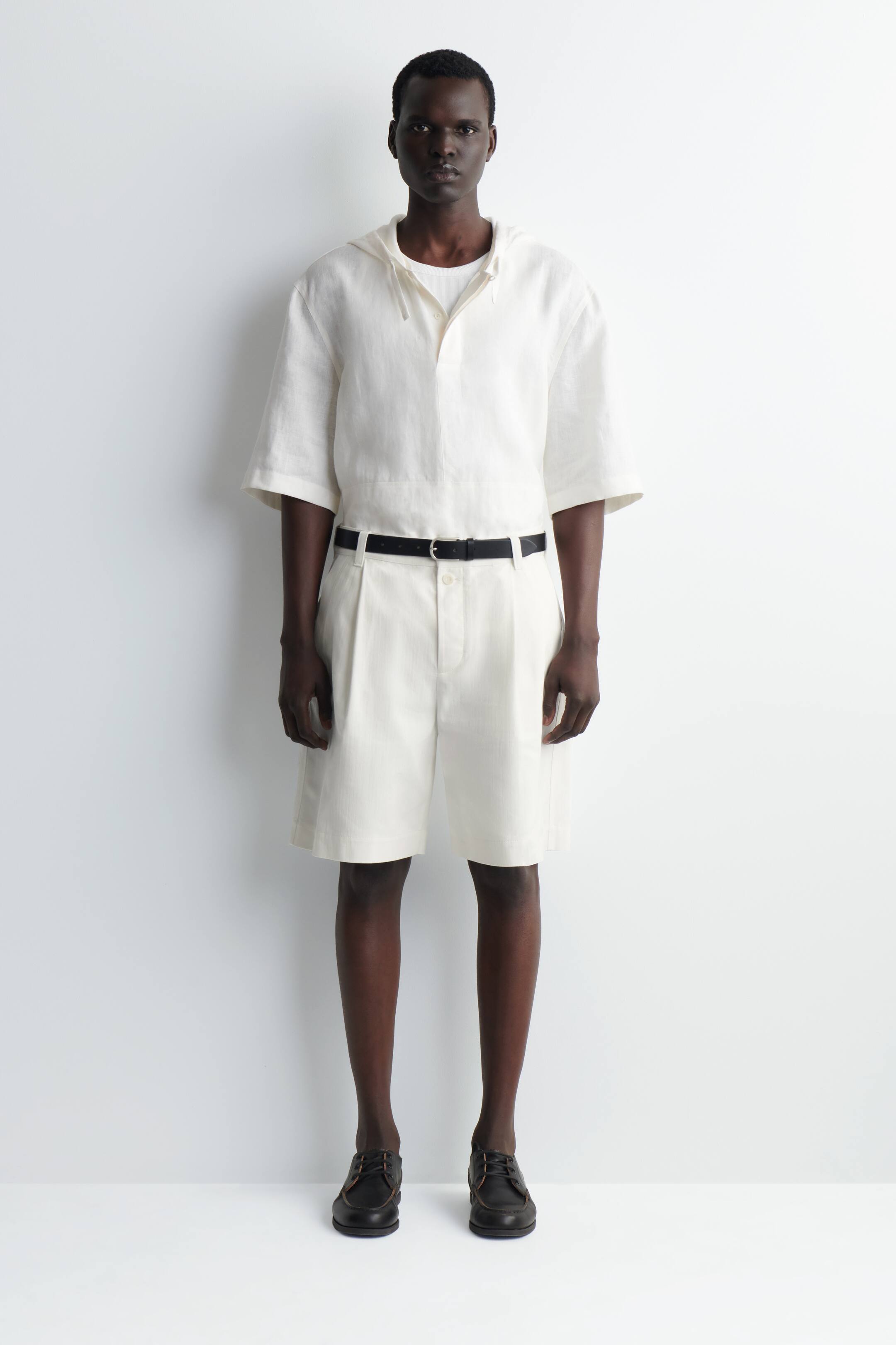 Visa större bild: Ett framåtvänd outfit består av en off-white kortärmad huvtröja, buren över en vit t-shirt, kombinerad med off-white plisserade shorts. Ett svart bälte markerar shortsen, och mörkbruna lädersnörskor fullbordar ensemblet.