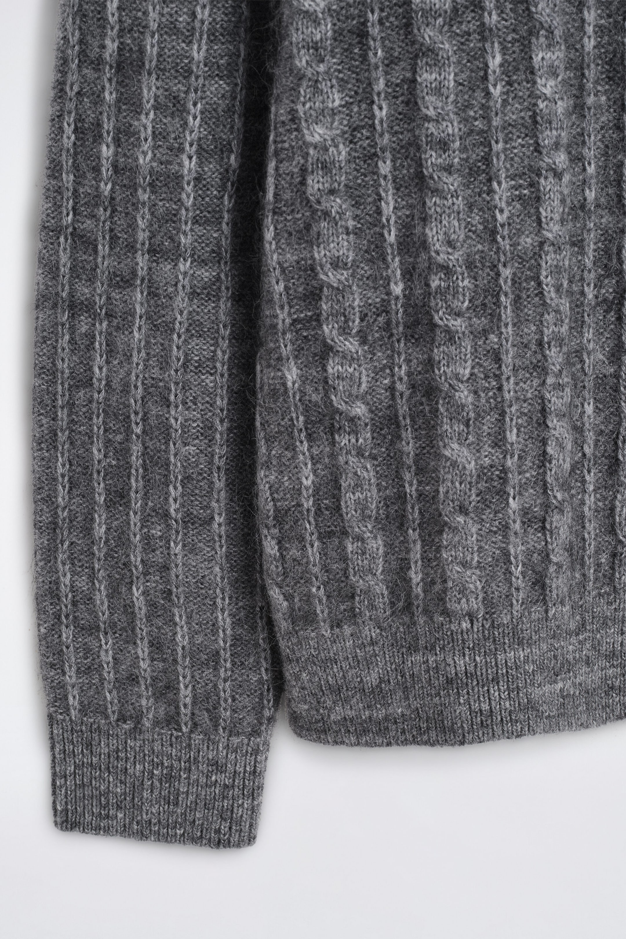 A grey cable-knit sweater is shown in a close-up front view, highlighting its textured knit construction. The sweater features a vertically ribbed pattern on the sleeve ending in a ribbed cuff, along with cable-knit detailing on the bodice that finishes with a ribbed hem. A grey cable-knit sweater is shown in a close-up front view, highlighting its textured knit construction. The sweater features a vertically ribbed pattern on the sleeve ending in a ribbed cuff, along with cable-knit detailing on the bodice that finishes with a ribbed hem.