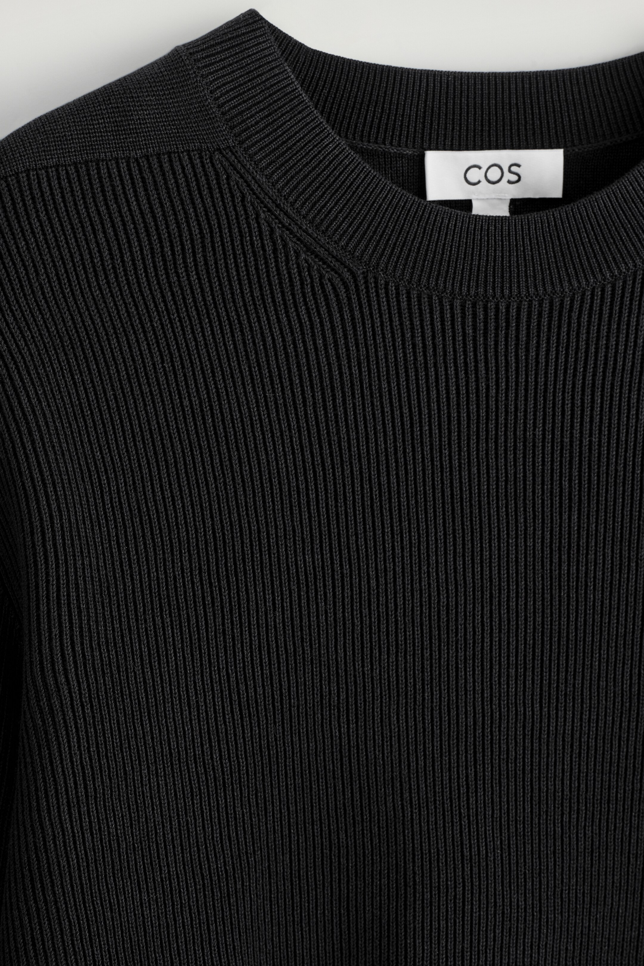 Agrandir l'image: Un pull noir côtelé est présenté de face, avec une texture côtelée verticale prononcée sur le corps. Il présente un col rond côtelé, et une étiquette de marque 'COS' blanche est visible à l'intérieur de l'encolure.