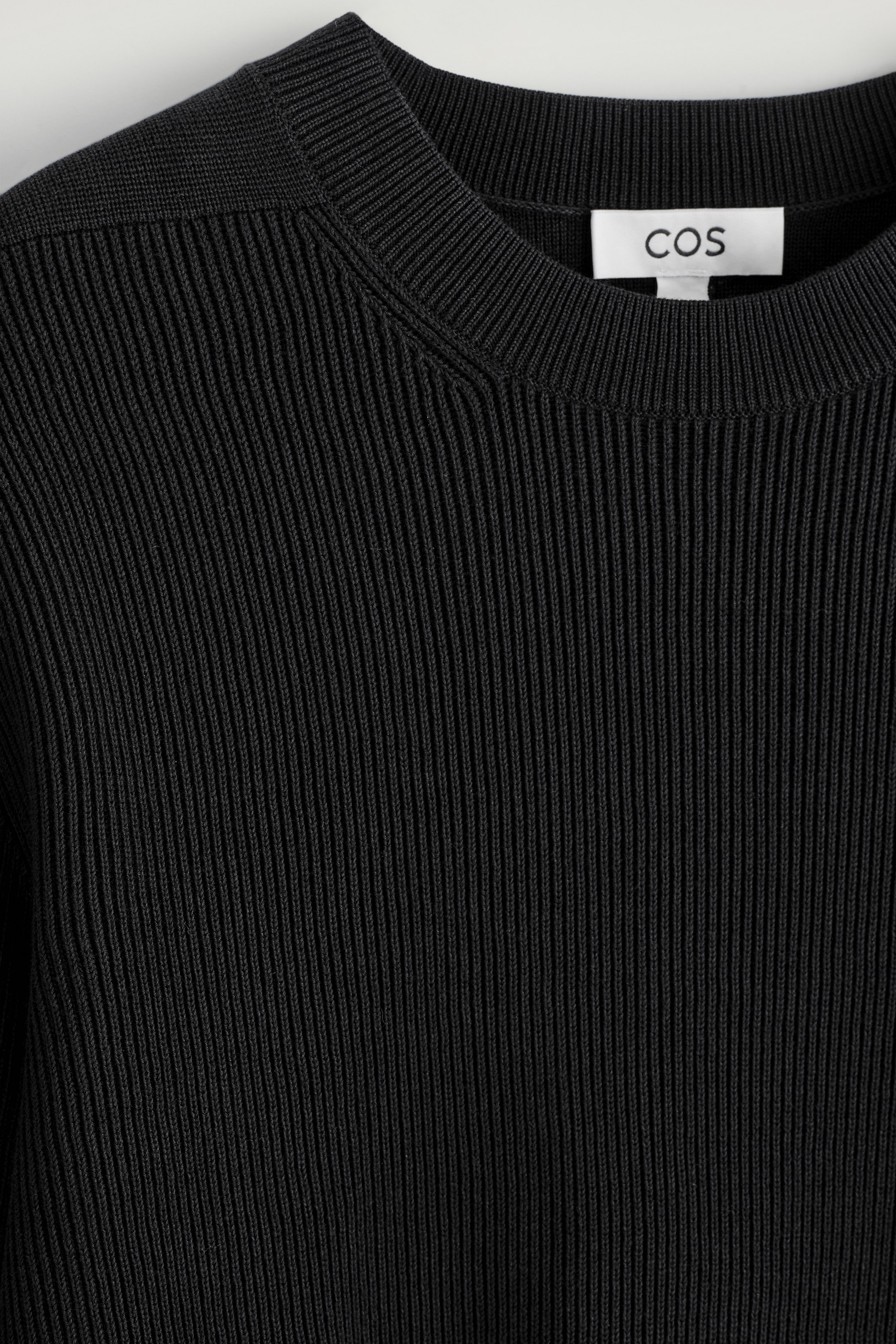 Agrandir l'image: Un pull noir côtelé est présenté de face, avec une texture côtelée verticale proéminente sur son corps. Il présente un col rond côtelé, et une étiquette de marque 'COS' blanche est visible à l'intérieur de l'encolure.