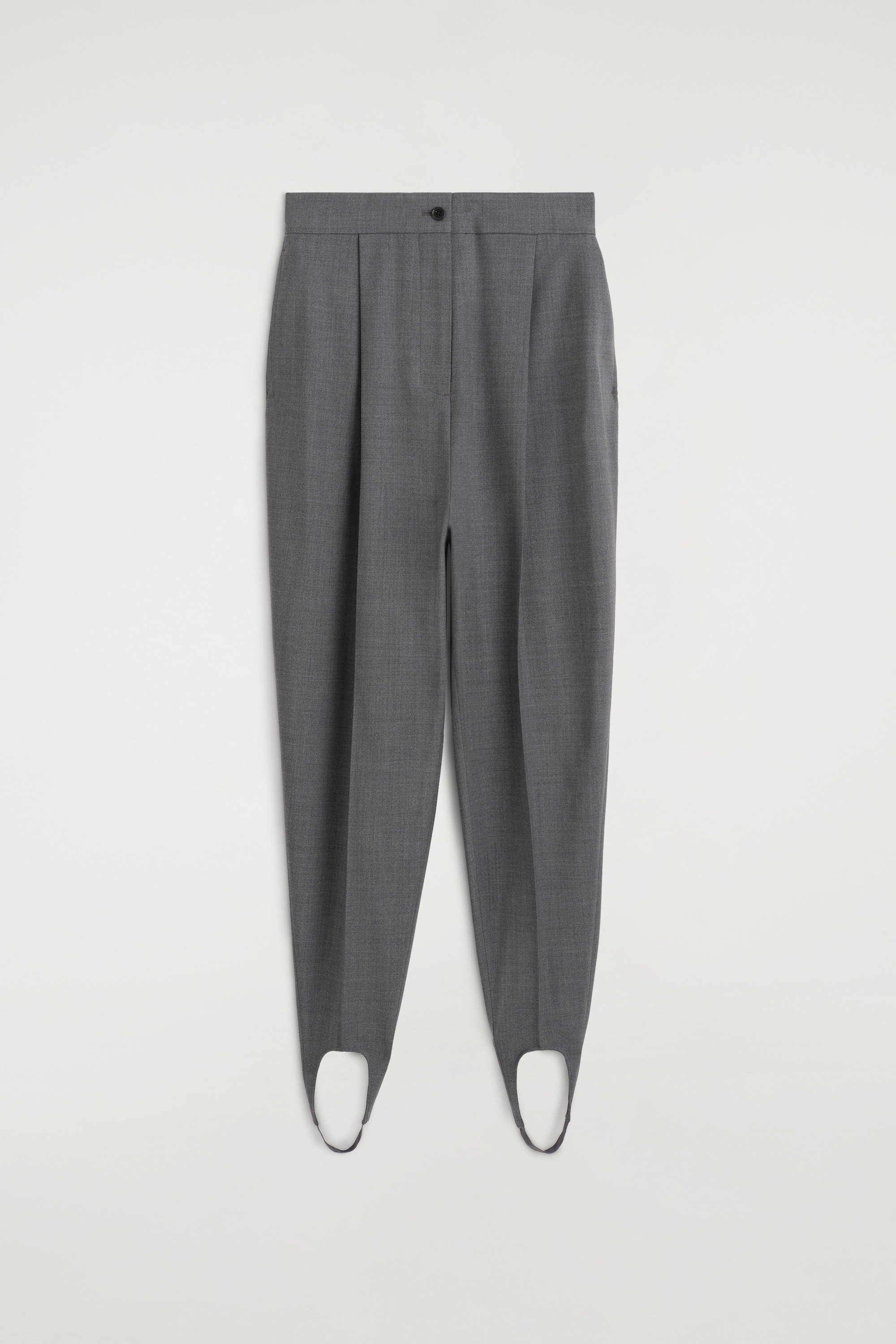 Questi pantaloni stirrup sartoriali grigio scuro presentano una vita alta con pieghe frontali stirate, chiusura con bottone e gamba affusolata. Le gambe terminano con staffe che passano sotto i talloni di mocassini neri, creando un caratteristico design cut-out alla caviglia.