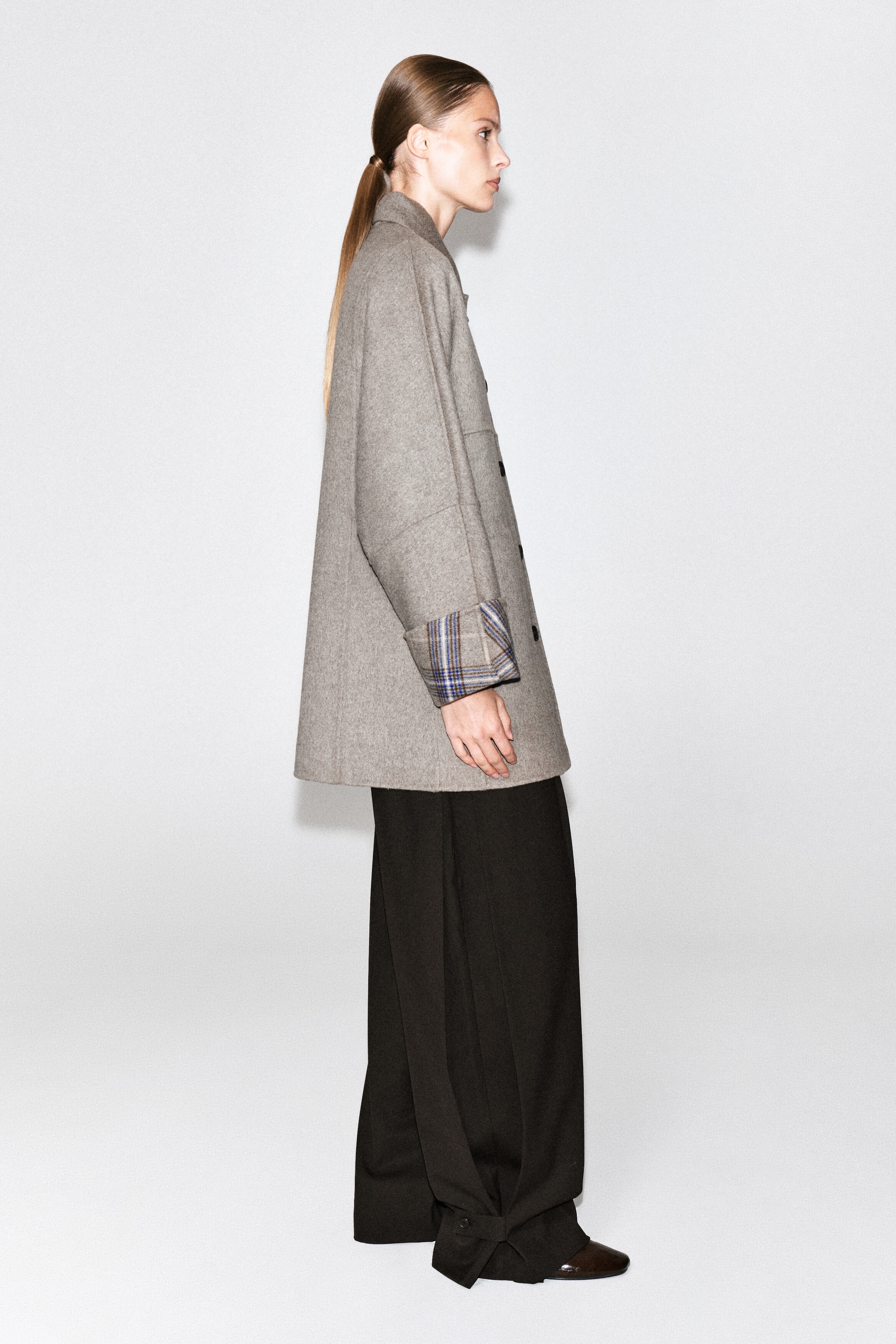 A beige double-faced coat is worn by the model standing in profile, paired with wide-leg black trousers. The mid-length coat features button closures and turned-up sleeve cuffs revealing a blue and brown check pattern on the reverse side.