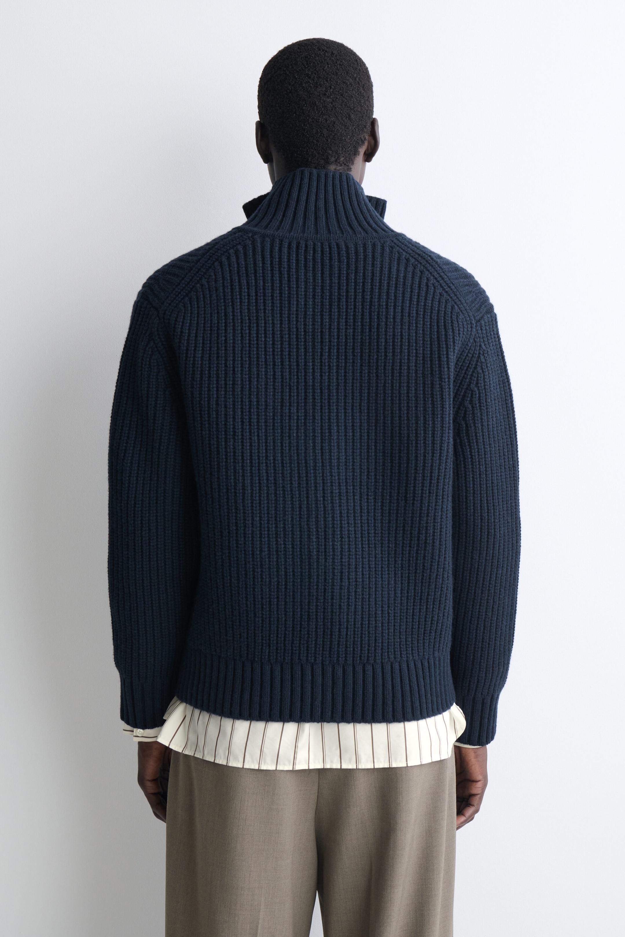 A navy blue ribbed-knit cardigan is shown from the back, featuring a high funnel neck, raglan sleeves, and a ribbed hem. The cardigan is layered over a cream-striped shirt and taupe trousers, showcasing its relaxed fit and richly textured knit. A navy blue ribbed-knit cardigan is shown from the back, featuring a high funnel neck, raglan sleeves, and a ribbed hem. The cardigan is layered over a cream-striped shirt and taupe trousers, showcasing its relaxed fit and richly textured knit.