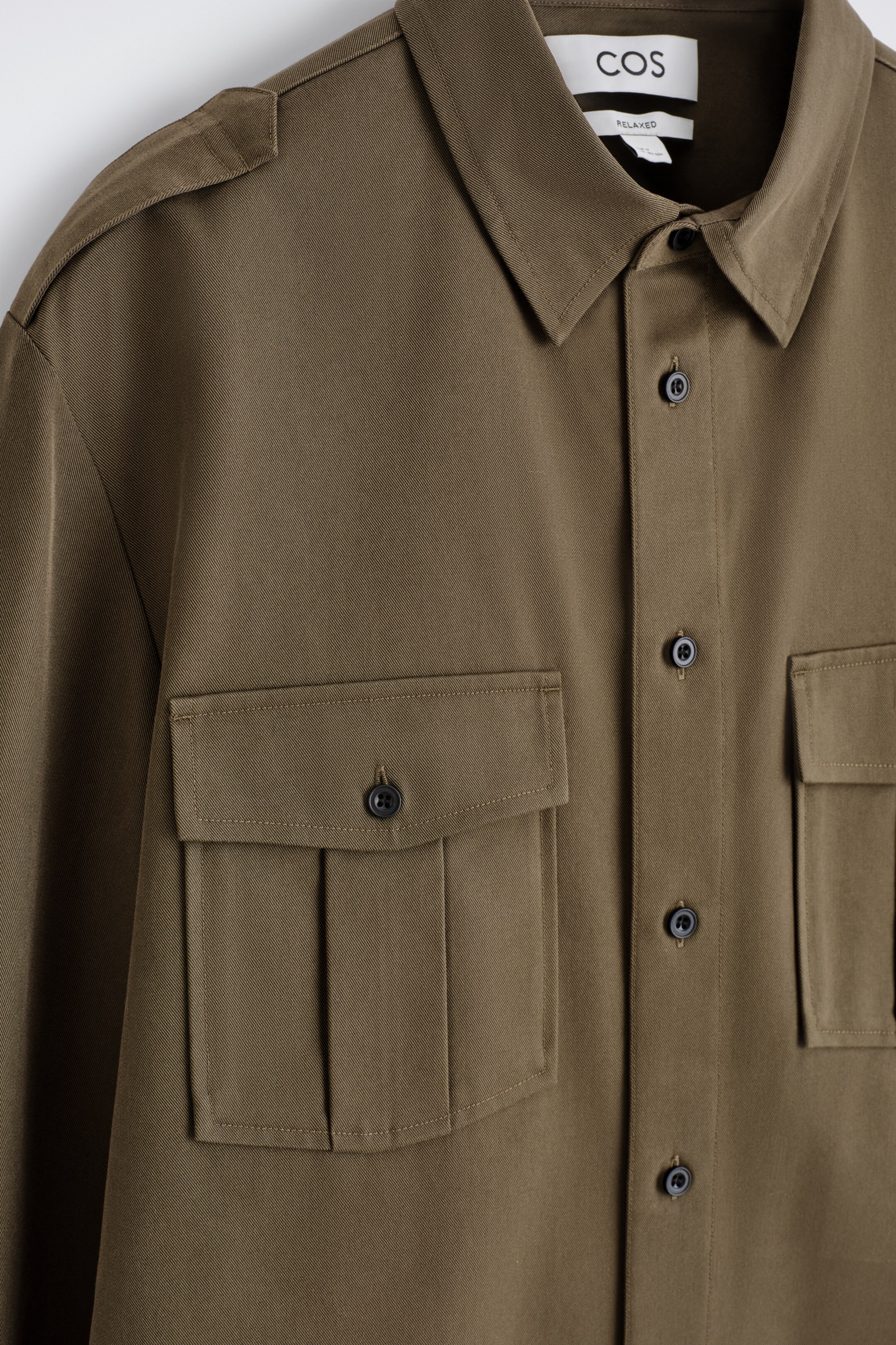 Ver imagen más grande: Se presenta una camisa de sarga verde oliva de FRENTE, con un cuello en punta, un frente abotonado con botones oscuros y charreteras en los hombros. Se pueden ver dos grandes bolsillos en el pecho con solapas y cierres de botón, con el bolsillo izquierdo también mostrando pliegues.
