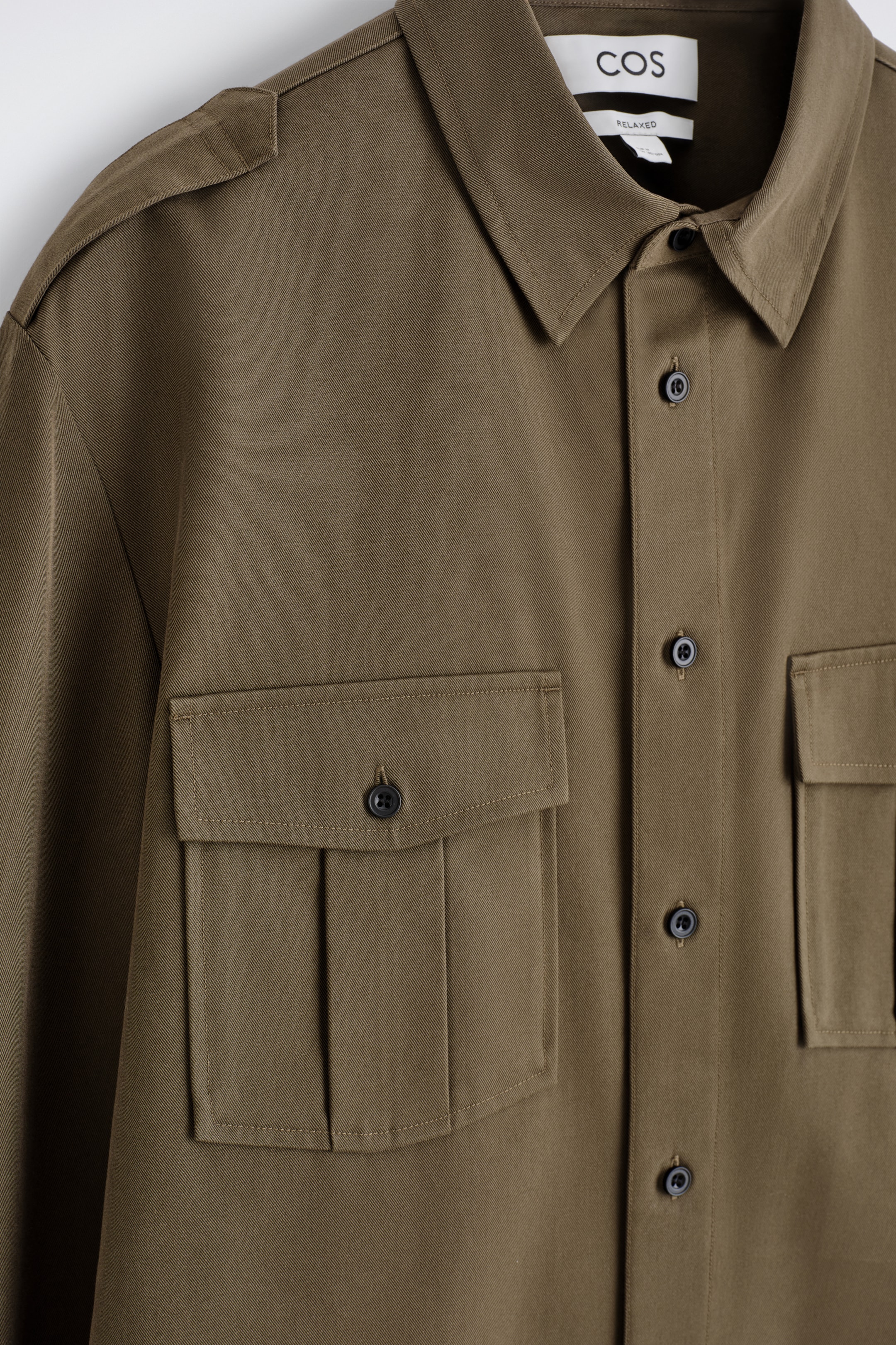 Agrandir l'image: Une chemise en sergé vert olive est présentée de FACE, avec un col pointu, une fermeture à boutons sur le devant avec des boutons foncés, et des épaulettes sur les épaules. Deux grandes poches poitrine avec rabat et fermetures à boutons sont visibles, la poche gauche présentant également des plis.