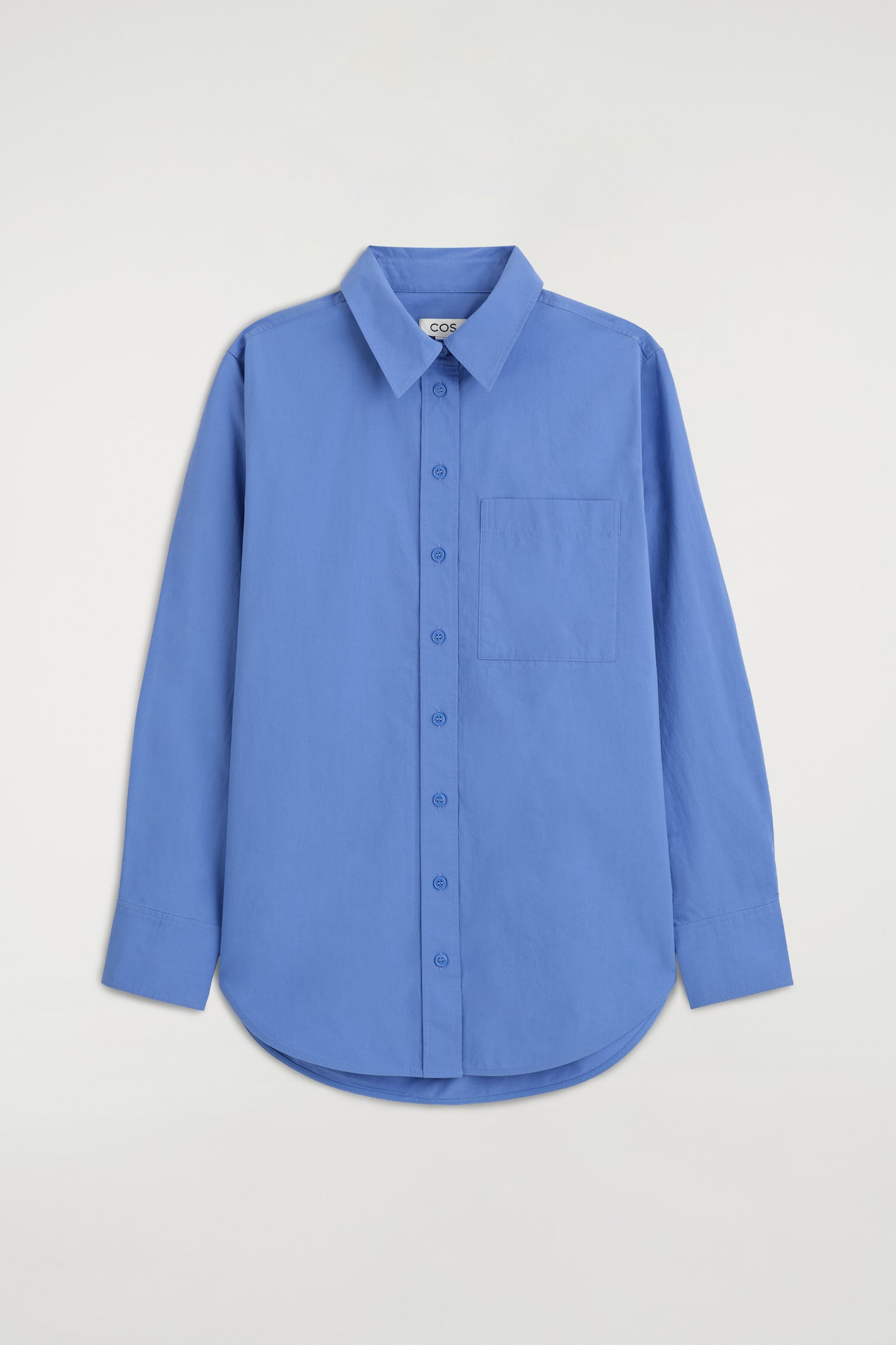 Uma camisa azul-cornflower de manga comprida e com botÃµes estÃ¡ virada para a frente, apresentando um colarinho clÃ¡ssico de pontas, uma carcela frontal com botÃµes azuis a condizer e um bolso de chapa no peito. A camisa exibe uma bainha curva e mangas compridas com punhos largos abotoados.
