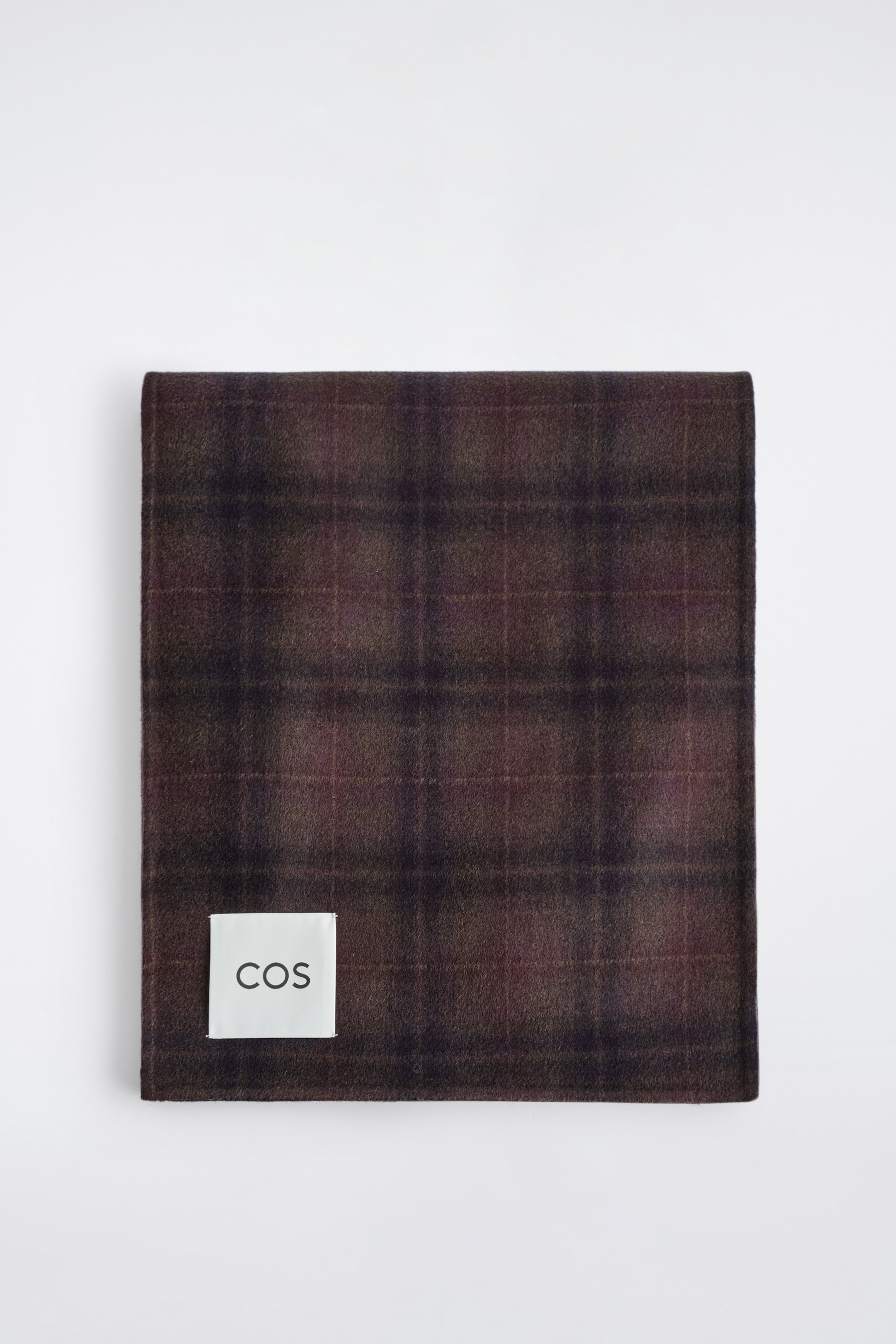 Agrandir l'image: Une écharpe tartan rectangulaire, présentée en vue de dessus, affiche un motif à carreaux bordeaux riche et une texture douce. Un patch logo rectangulaire blanc avec "COS" en texte noir est visible dans le coin inférieur gauche.