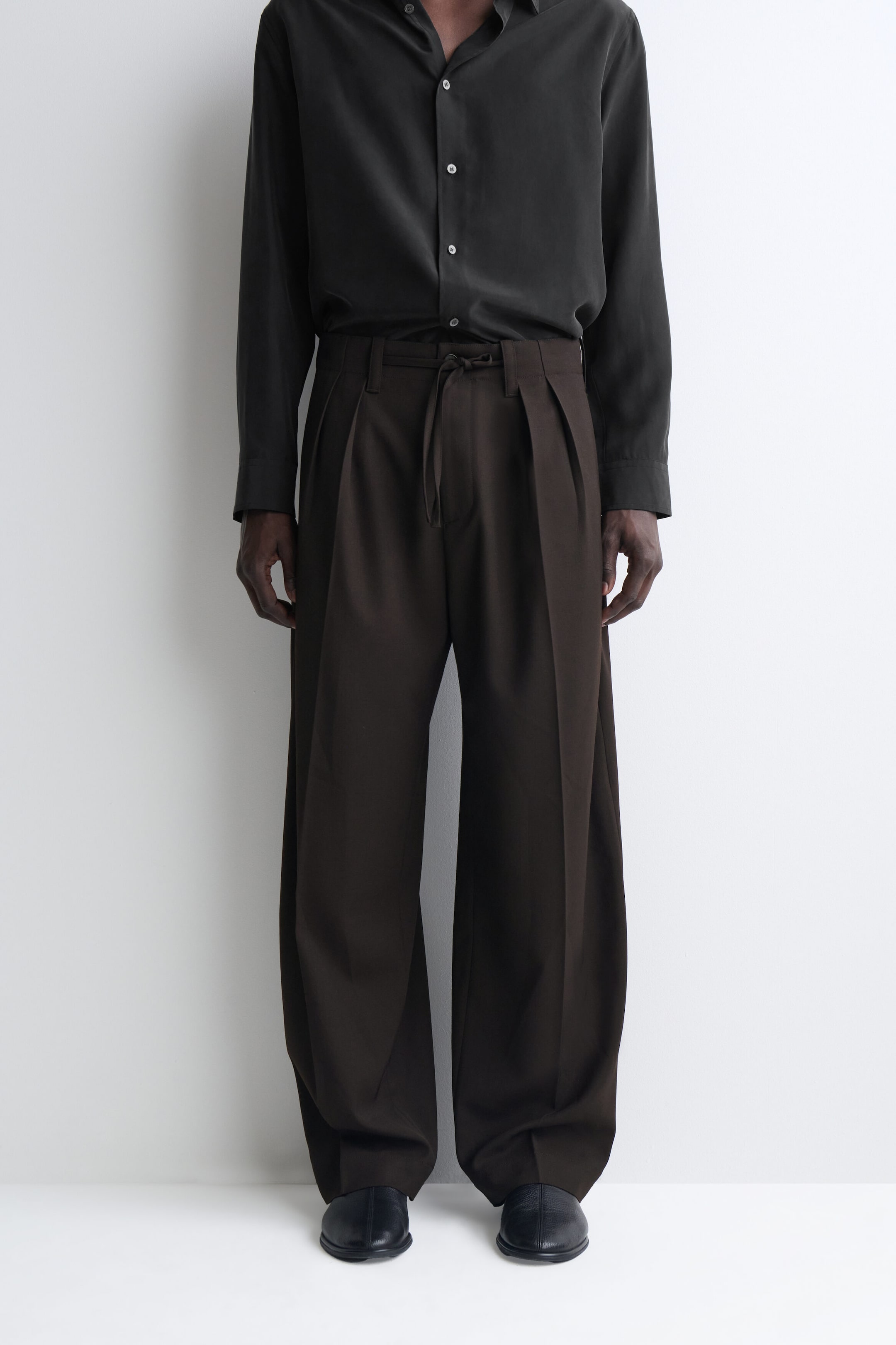These dark brown wide-leg trousers feature double front pleats, belt loops, and a thin drawstring tie at the waist. The hems drape in a relaxed fit down to the ankles and are styled with a tucked-in dark long-sleeve shirt. These dark brown wide-leg trousers feature double front pleats, belt loops, and a thin drawstring tie at the waist. The hems drape in a relaxed fit down to the ankles and are styled with a tucked-in dark long-sleeve shirt.
