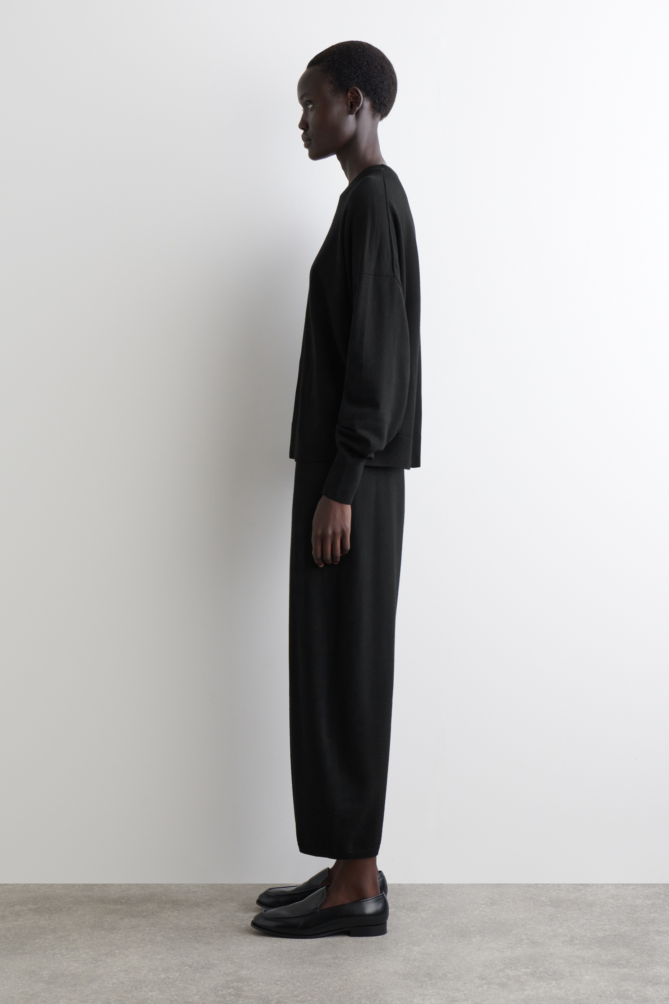 Ver imagen más grande: Un conjunto de punto negro de dos piezas se presenta de perfil lateral. La parte superior presenta un ajuste holgado con mangas largas y relajadas y puños, mientras que los pantalones coordinados muestran una silueta de pierna recta que termina justo por encima del tobillo.