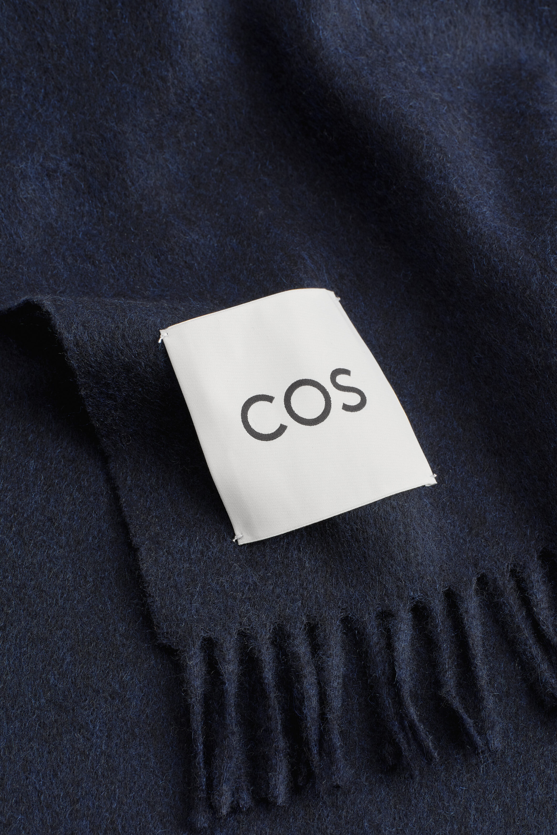 A dark navy cashmere scarf displays a soft, brushed texture and a fringed hem, with a white rectangular brand label reading "COS" stitched near the edge. A dark navy cashmere scarf displays a soft, brushed texture and a fringed hem, with a white rectangular brand label reading "COS" stitched near the edge.