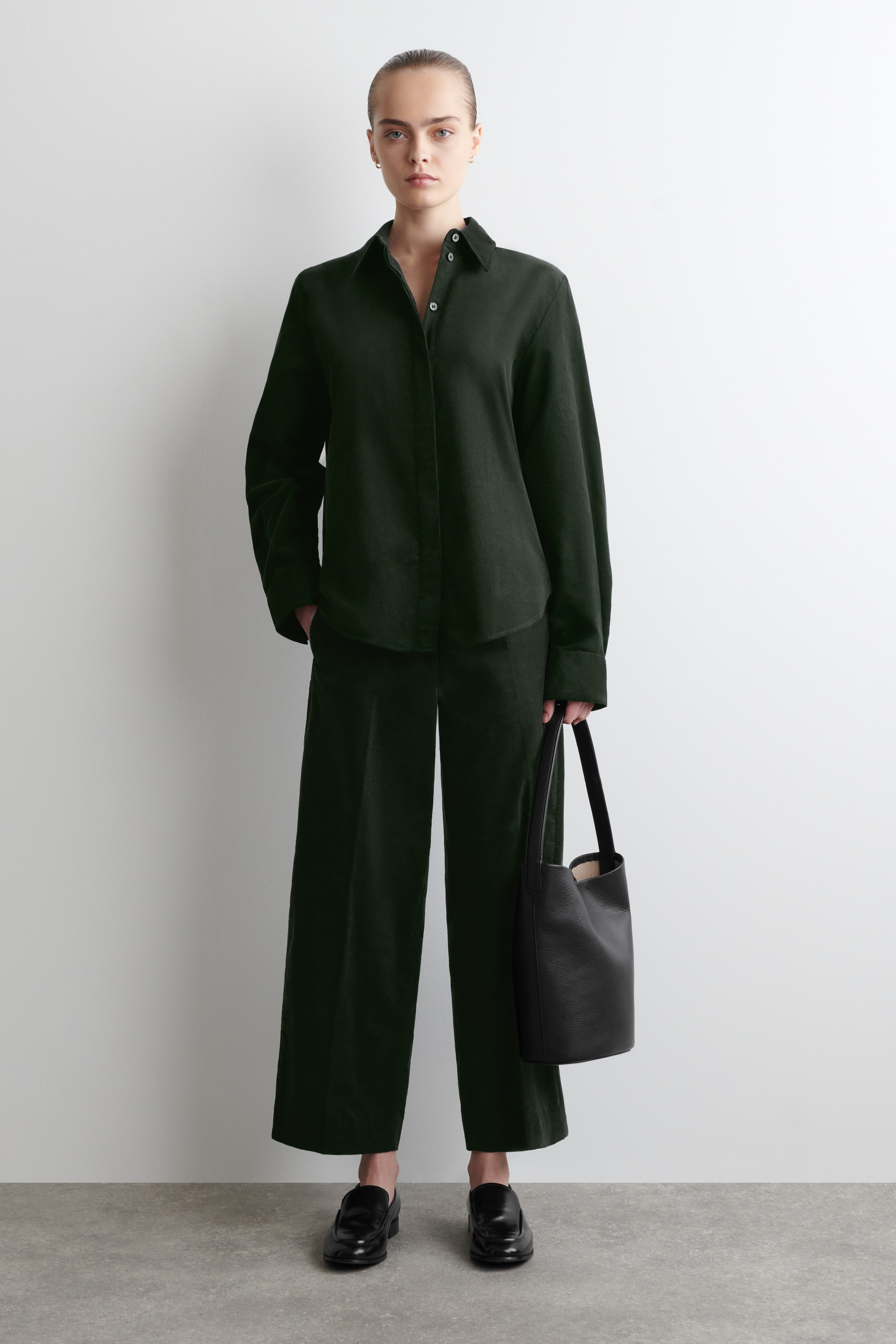 Visualizza immagine più grande: Una camicia verde scuro a maniche lunghe con bottoni, colletto a punta e orlo arrotondato viene mostrata con pantaloni a gamba larga coordinati, presentando una vista frontale completa. Una borsa a secchiello in pelle nera viene tenuta in mano e mocassini neri vengono indossati, completando l'ensemble.