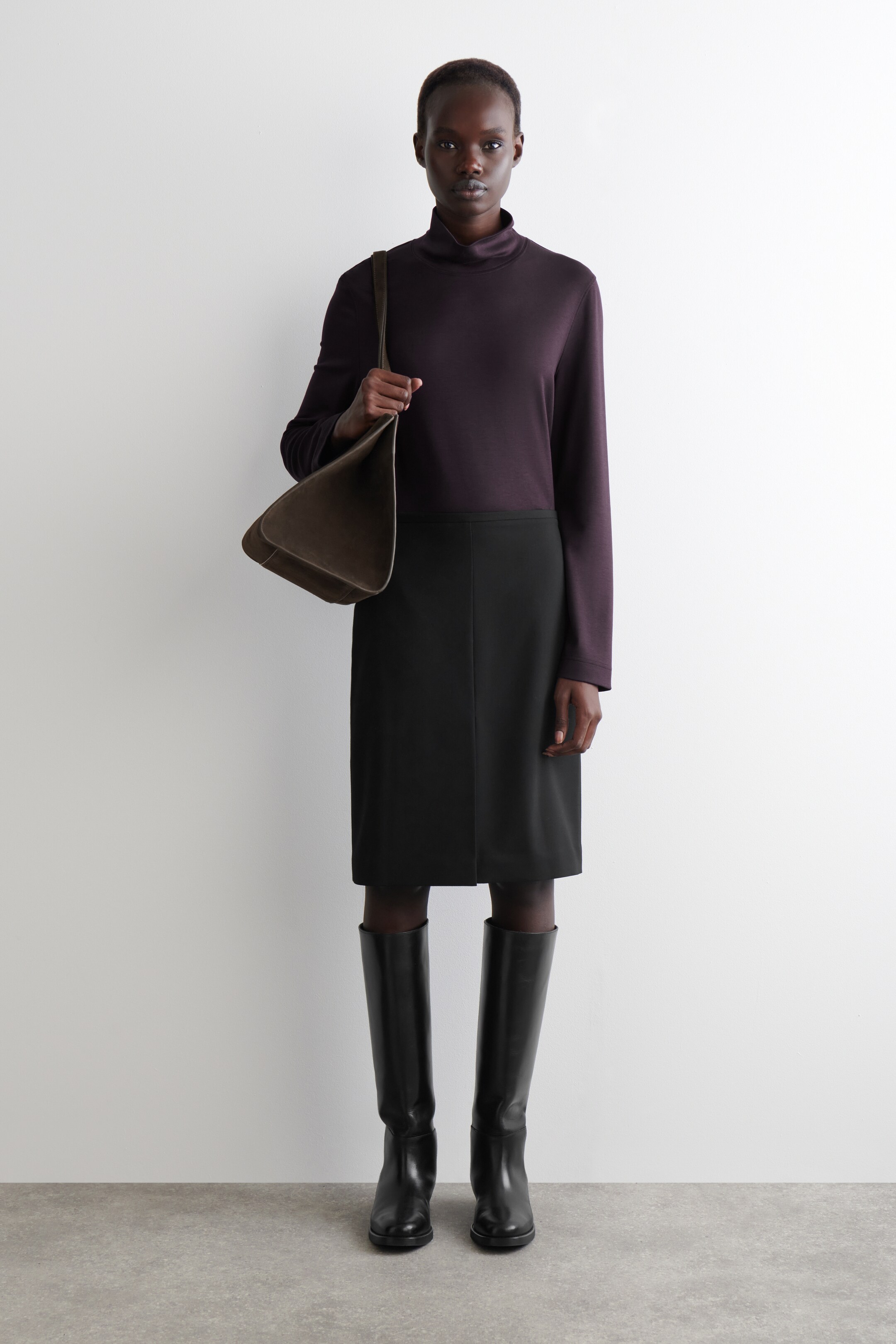 Agrandir l'image: Un haut à col montant à manches longues couleur aubergine foncée est porté avec une jupe crayon noire longueur genou et des bottes plates noires hauteur genou. Un sac à bandoulière non structuré couleur olive foncée est tenu dans la main droite, avec sa sangle drapée sur l'épaule.