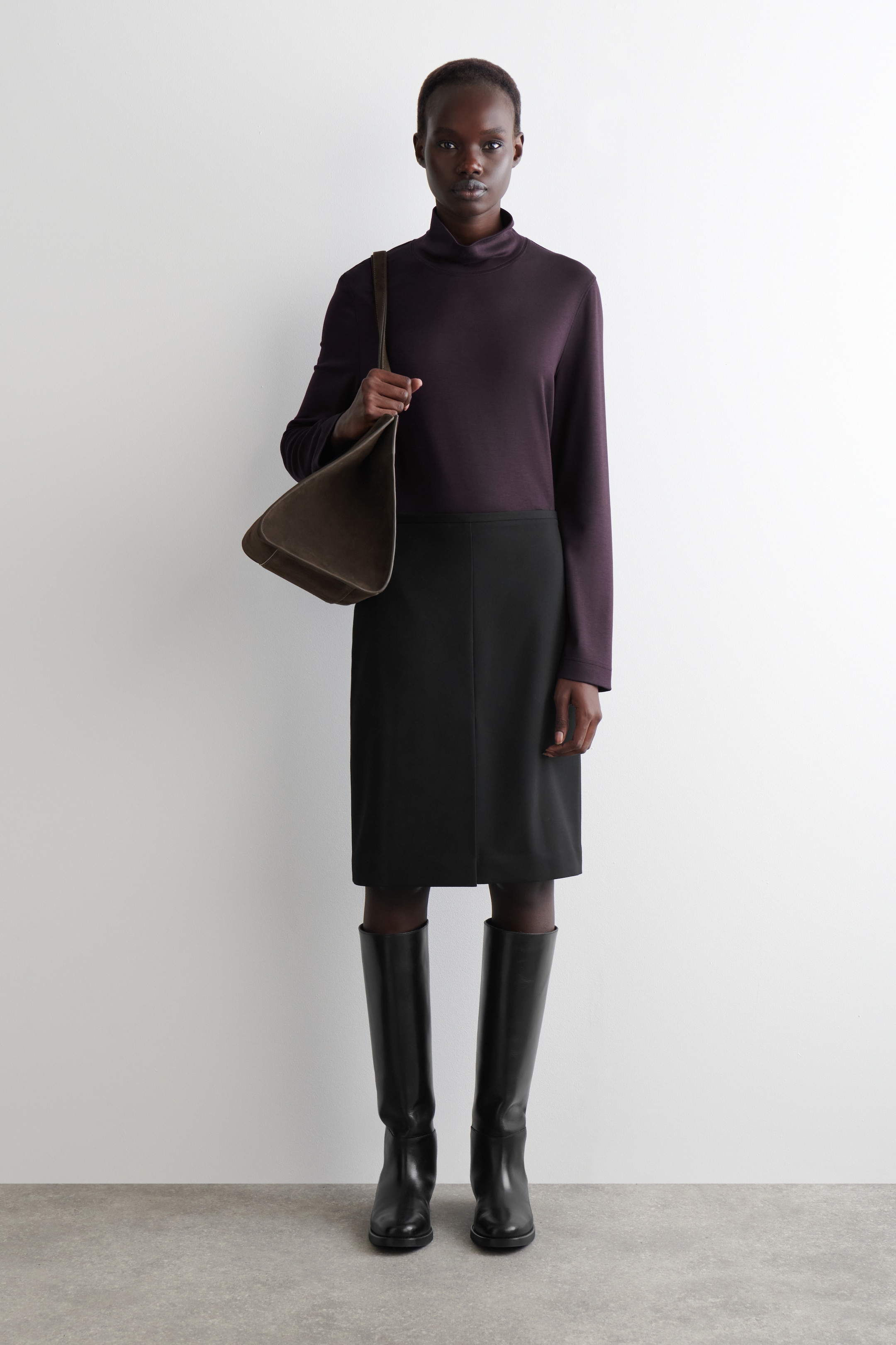 Agrandir l'image: Un haut à col montant à manches longues couleur aubergine foncé est porté avec une jupe crayon noire longueur genou et des bottes plates noires hauteur genou. Un sac à bandoulière non structuré couleur olive foncé est tenu dans la main droite, avec sa sangle drapée sur l'épaule.