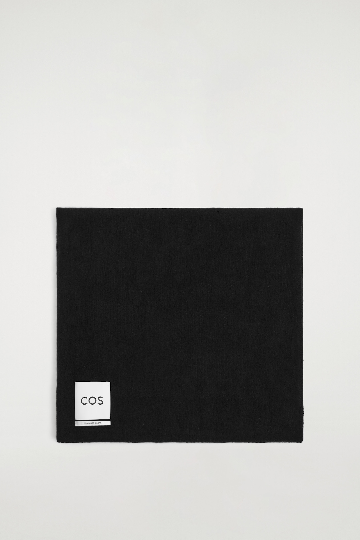 A black cashmere scarf is laid flat and folded into a neat square, showcasing its soft texture. A white rectangular label featuring the black text "COS" and "100% CASHMERE" appears in the bottom-left corner.