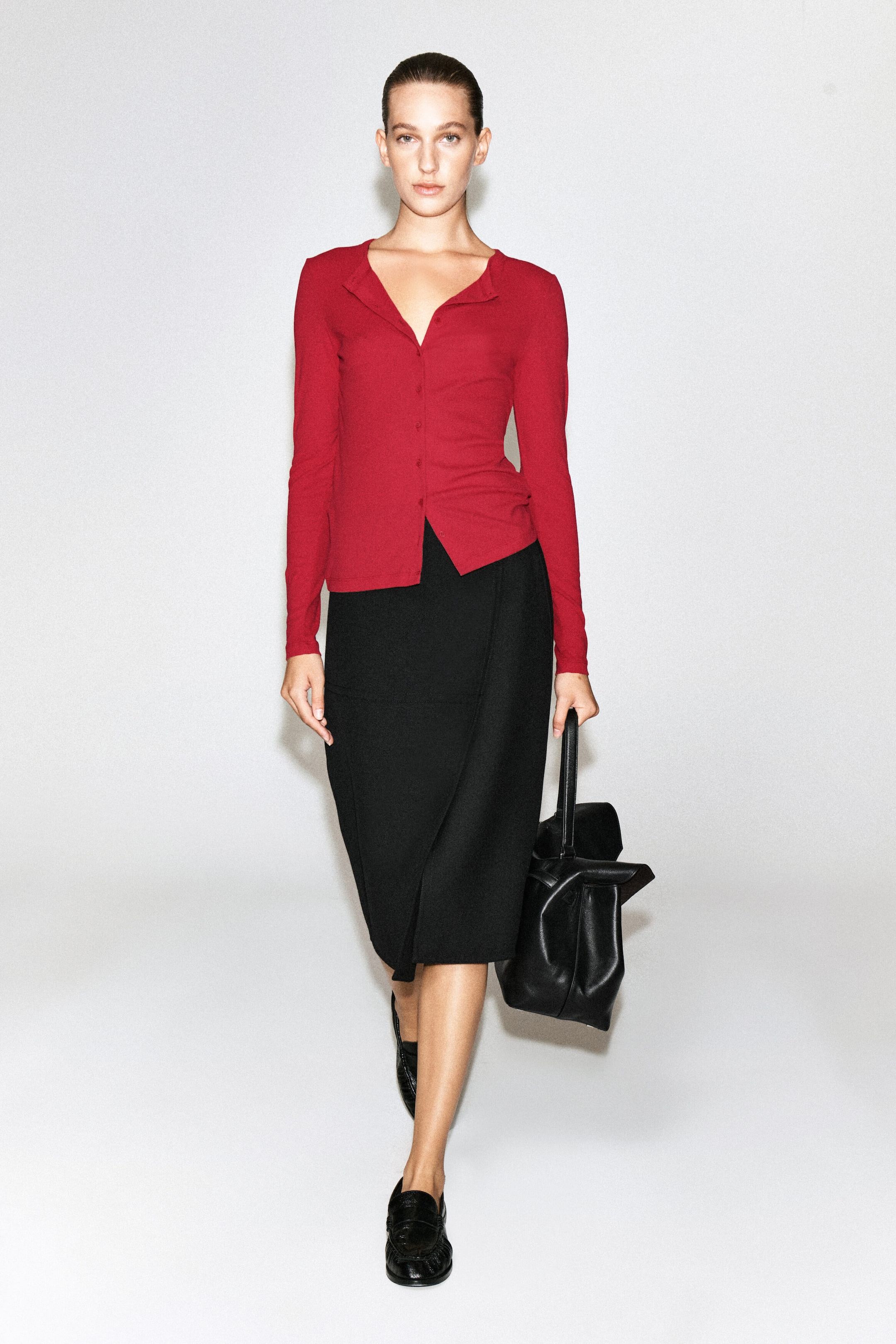 The model is wearing a red jersey cardigan top featuring long sleeves, a button-front closure, and an open collar. The top is styled with a black midi skirt, black loafers, and a black leather handbag carried by the model. The model is wearing a red jersey cardigan top featuring long sleeves, a button-front closure, and an open collar. The top is styled with a black midi skirt, black loafers, and a black leather handbag carried by the model.