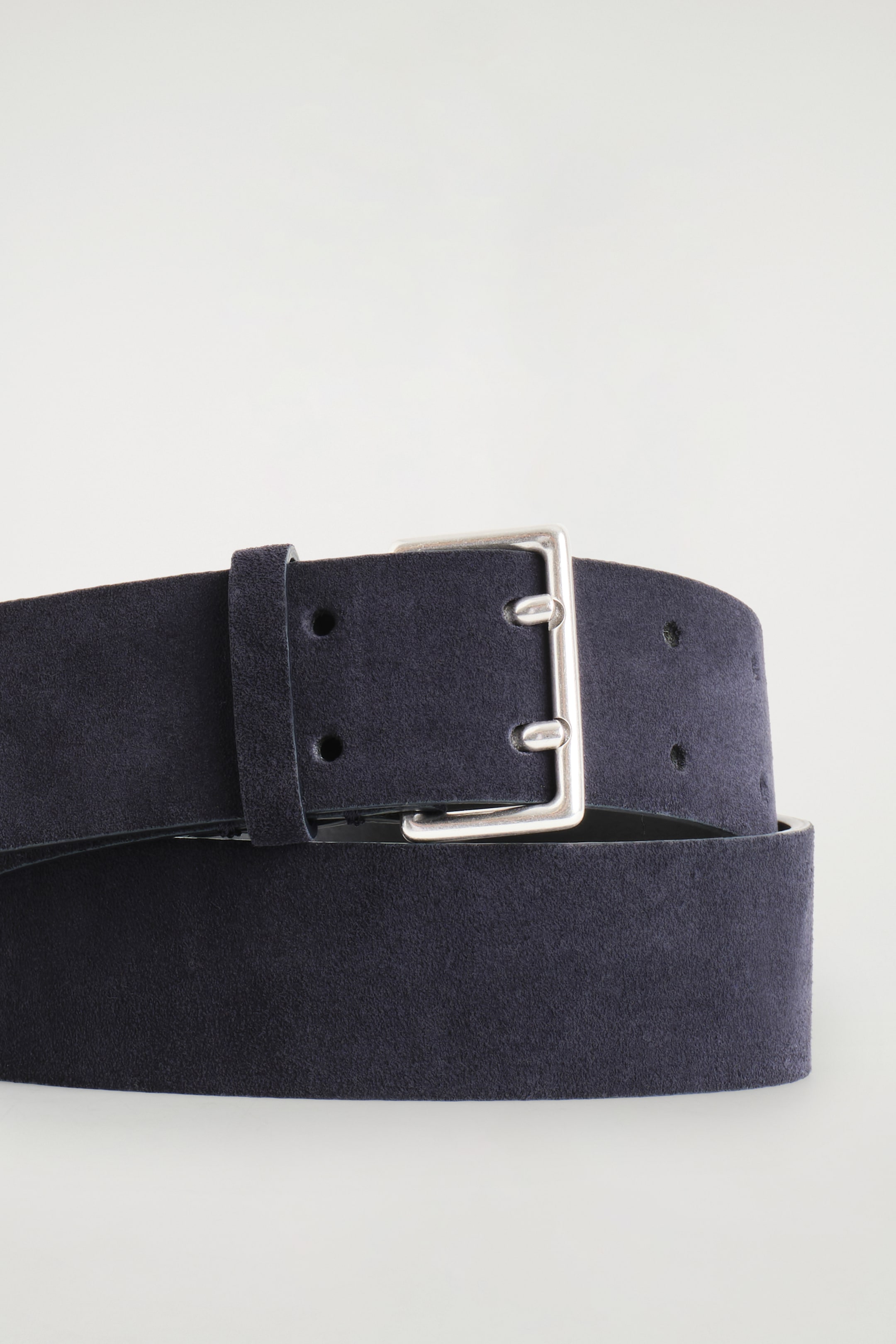 View larger image: A wide, dark navy suede belt is being presented from a low angle, featuring a squared silver-tone double-prong buckle. The belt strap is showing a softly brushed texture, with a matching belt loop and visible eyelets along its length.