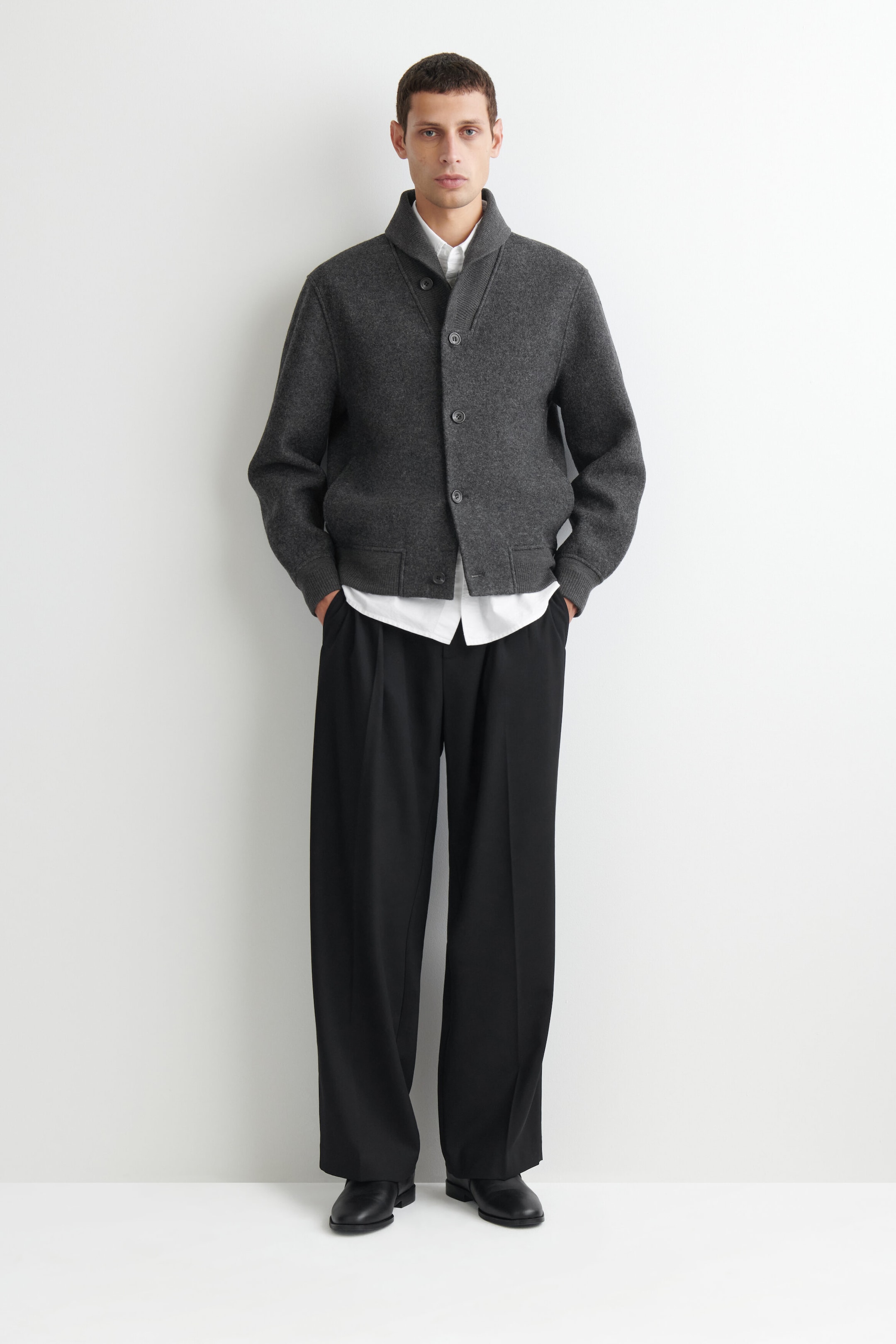 The boiled wool shawl-collar jacket is shown from the front, featuring a button-front closure, a thick shawl collar, and ribbed knit trim at the cuffs and hem. The jacket is layered over a white collared shirt and paired with black trousers, highlighting its relaxed fit and cropped hemline. The boiled wool shawl-collar jacket is shown from the front, featuring a button-front closure, a thick shawl collar, and ribbed knit trim at the cuffs and hem. The jacket is layered over a white collared shirt and paired with black trousers, highlighting its relaxed fit and cropped hemline.