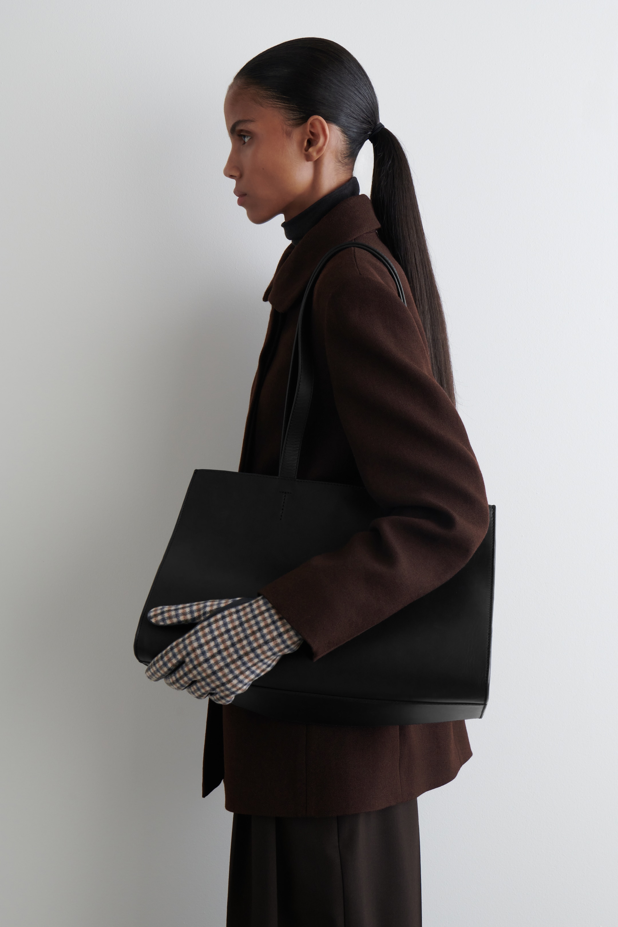 View larger image: A hand, positioned facing left, is wearing a patterned glove and holding a black tote bag. The glove features a small, tight brown, beige, and dark blue gingham check pattern, fitting snugly on the hand.