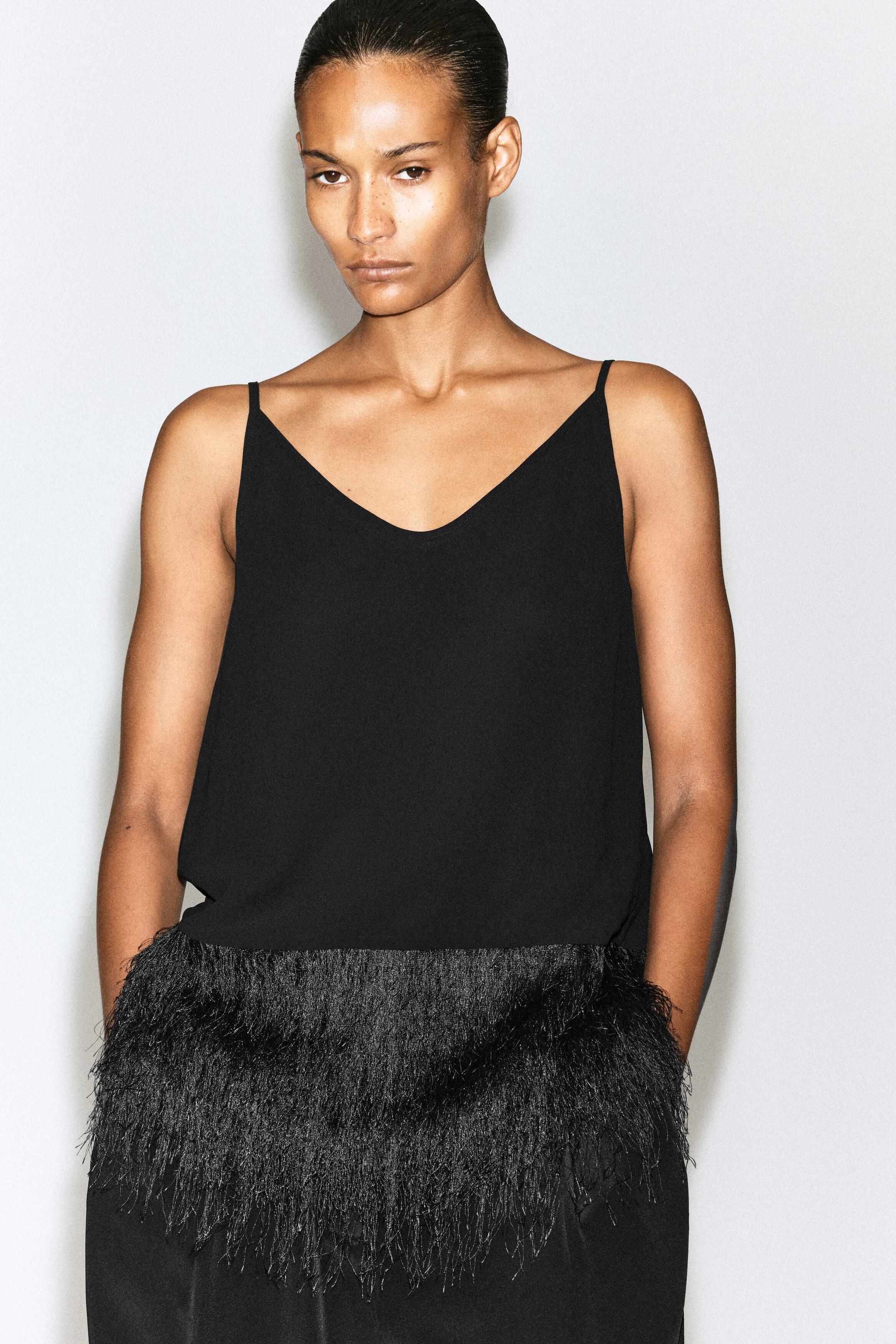 En svart camisole-topp med tunna axelband, rundad halsringning och en bred kant av luftig frans vid fållen bärs av modellen. Modellen matchar toppen med svarta byxor i samma stil. En svart camisole-topp med tunna axelband, rundad halsringning och en bred kant av luftig frans vid fållen bärs av modellen. Modellen matchar toppen med svarta byxor i samma stil.