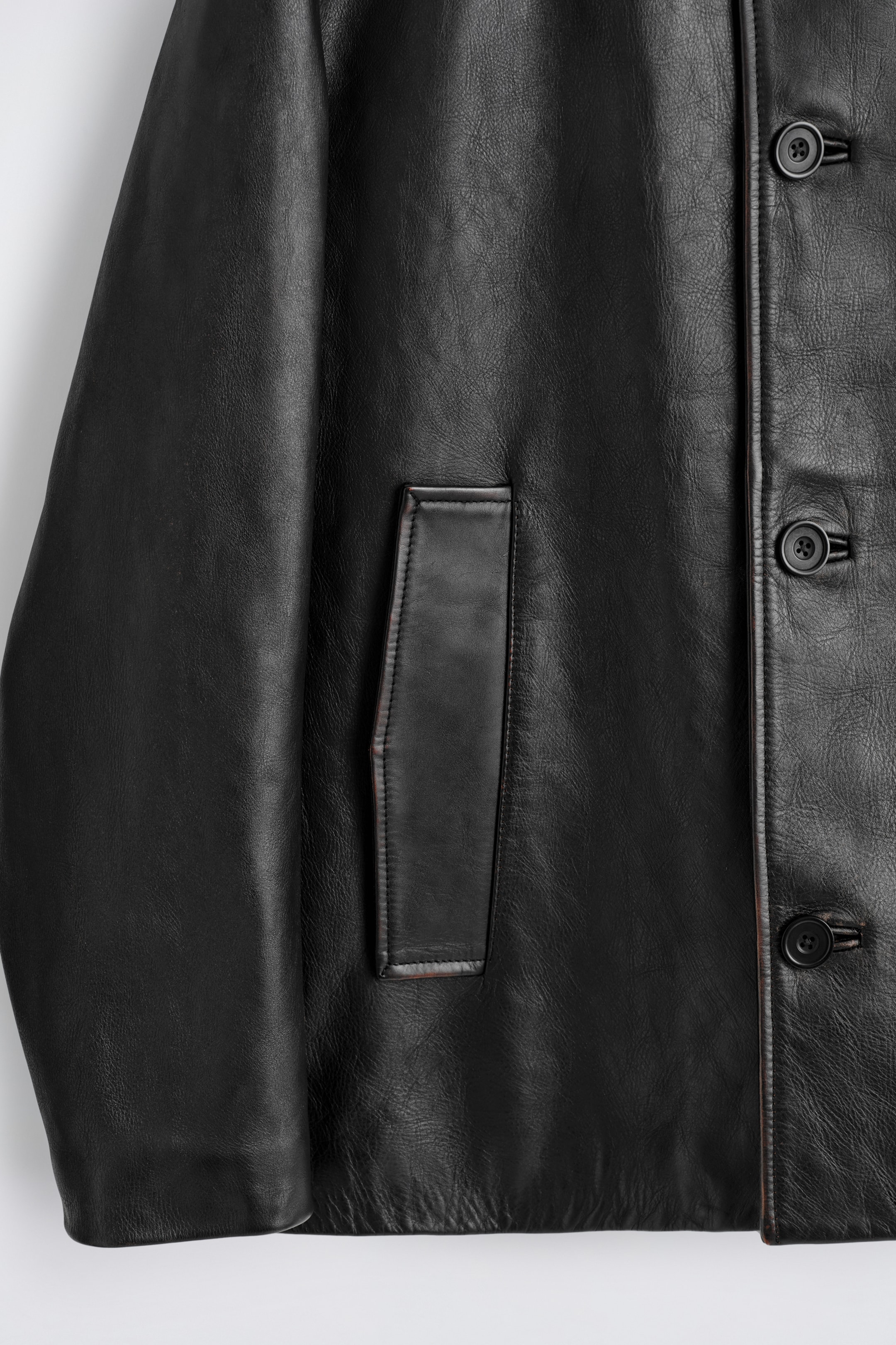 Agrandir l'image: Une veste en cuir noir présente son devant, avec une patte de boutonnage ornée de boutons ronds noirs. Une poche passepoilée horizontale apparaît sur le côté gauche de la veste, et des nuances brun-rouge accentuent les bords du cuir et l'ouverture de la poche.