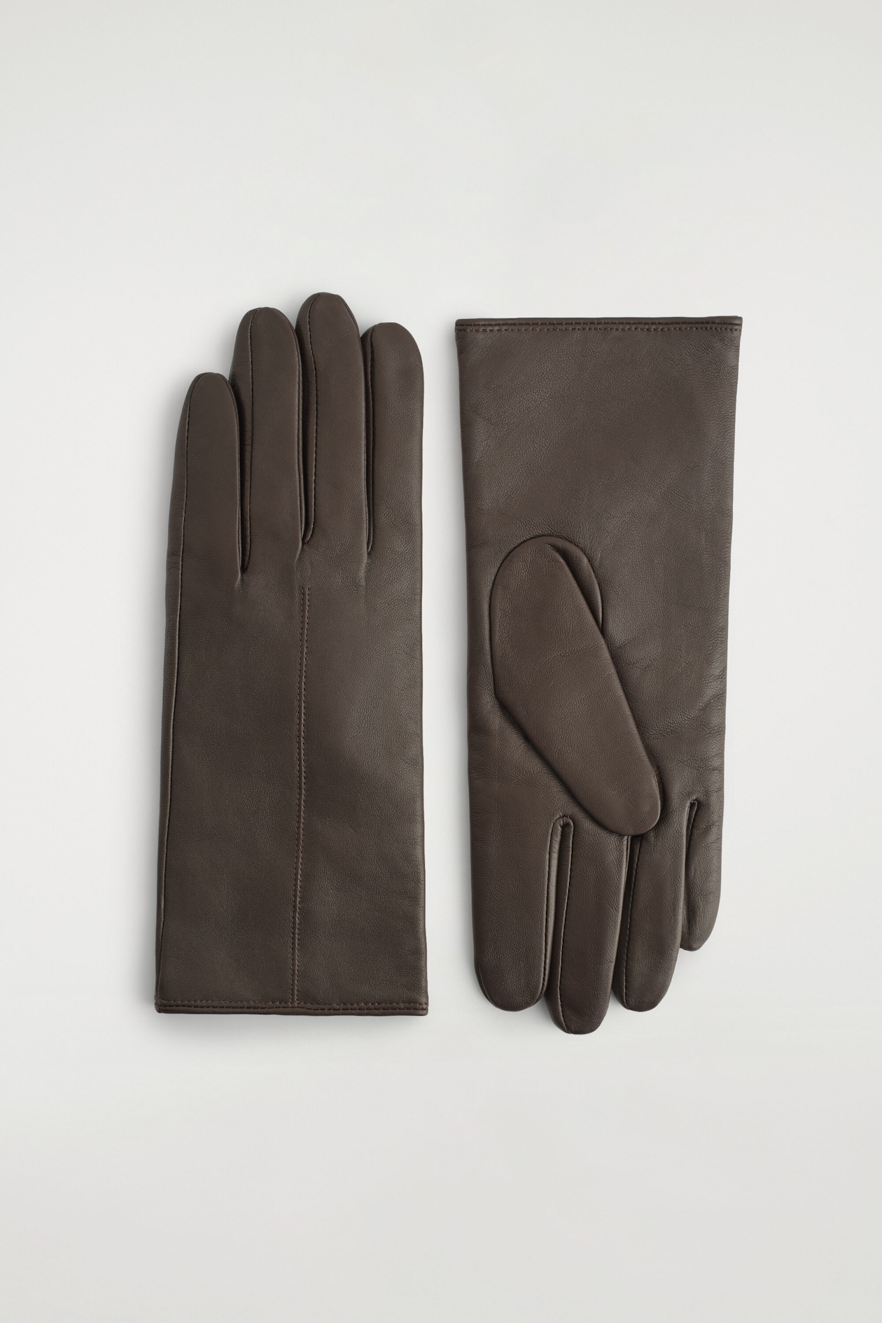 Une paire de gants en cuir brun foncé est posée à plat, mettant en valeur les vues du dos et de la paume. Le gant de gauche présente une seule couture verticale sur le dos de la main, tandis que le gant de droite montre le côté paume avec un détail de pouce cousu.