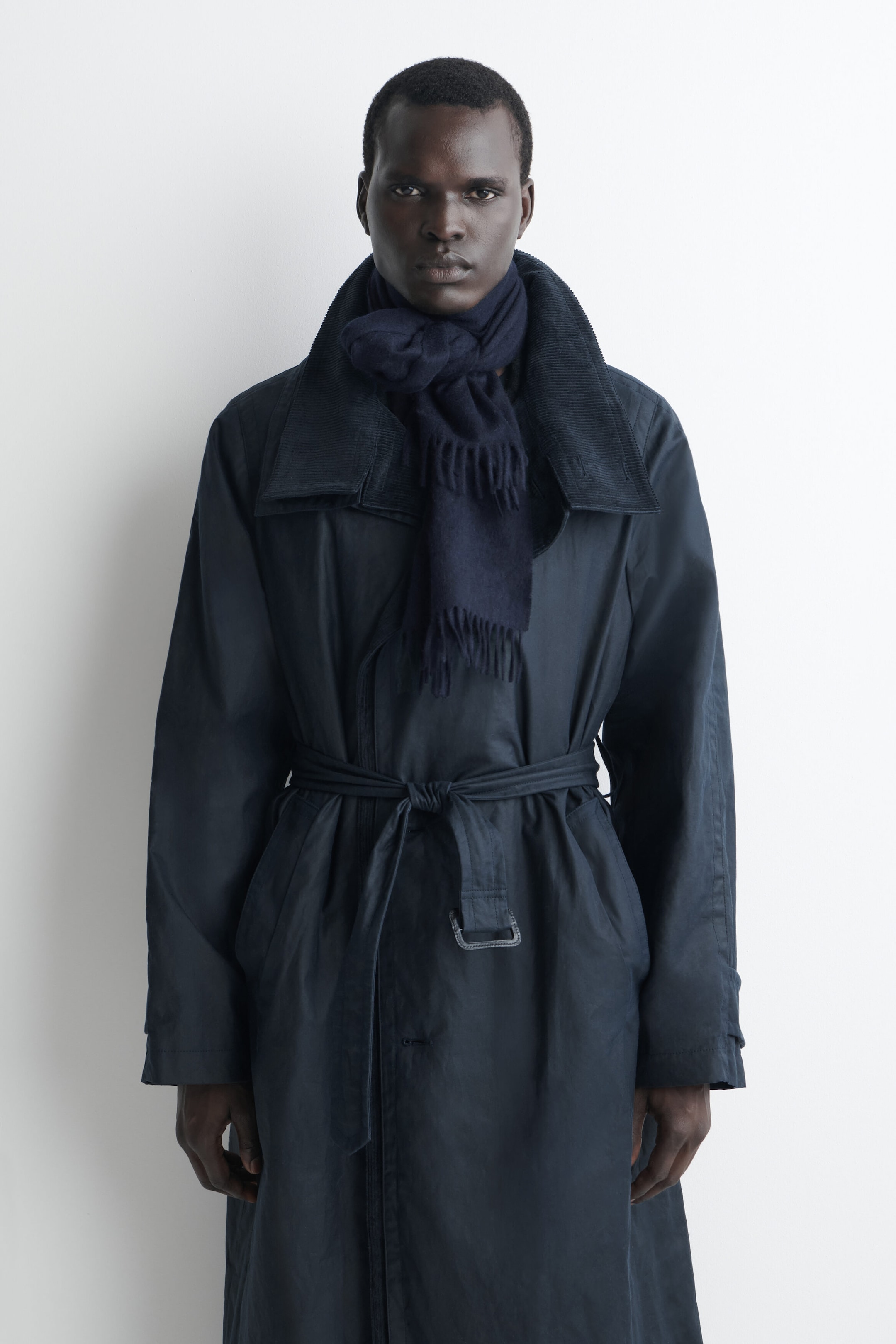 A navy blue fringed cashmere scarf is shown in a front-facing view, worn knotted around the neck with its fringed ends hanging down the chest. The scarf is layered inside the collar of a dark belted trench coat, highlighting its soft woven texture. A navy blue fringed cashmere scarf is shown in a front-facing view, worn knotted around the neck with its fringed ends hanging down the chest. The scarf is layered inside the collar of a dark belted trench coat, highlighting its soft woven texture.