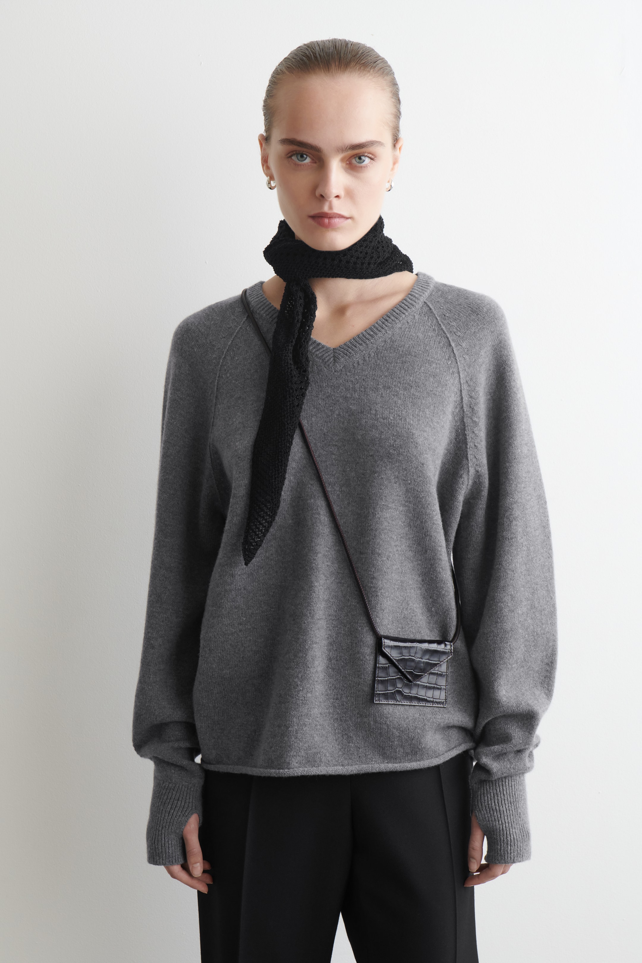 Grösseres Bild ansehen: Ein grauer V-Ausschnitt-Strickpullover wird von vorne präsentiert, mit langen Raglanärmeln, gerippten Bündchen mit integrierten Daumenlöchern und einem gerippten Saum. Ein schwarzer gehäkelter Schal wird um den V-Ausschnitt getragen, und eine kleine schwarze Umhängetasche mit Krokodilprägung hängt über dem Pullover.