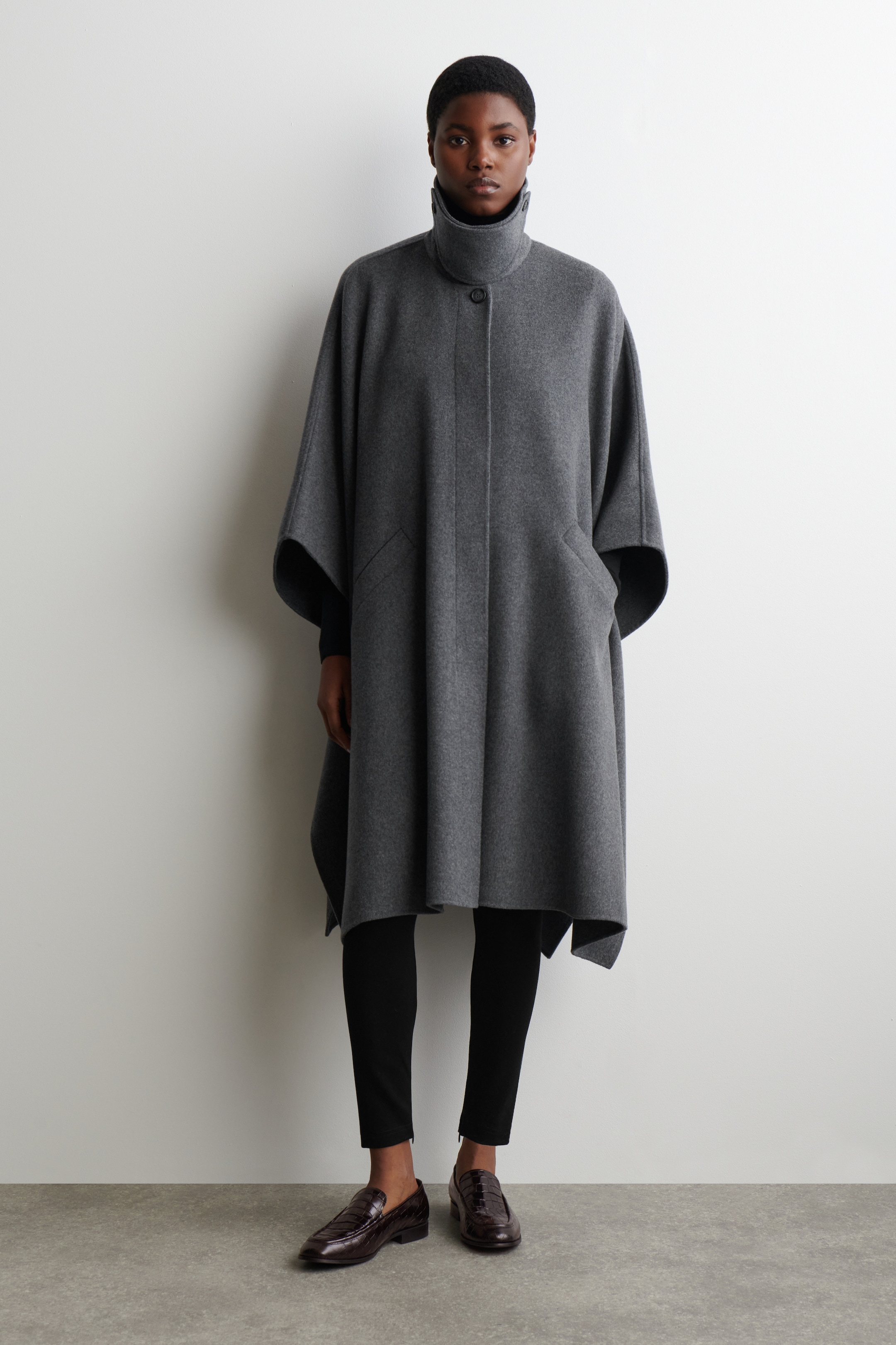 Agrandir l'image: Une cape en laine feutrée grise est présentée de face. Elle présente un col droit haut avec une fermeture à boutons visible, une patte de boutonnage dissimulée à l'avant et deux poches à ouverture en biais sur le devant, tout en drapant avec de larges ouvertures pour les bras et un ourlet asymétrique.