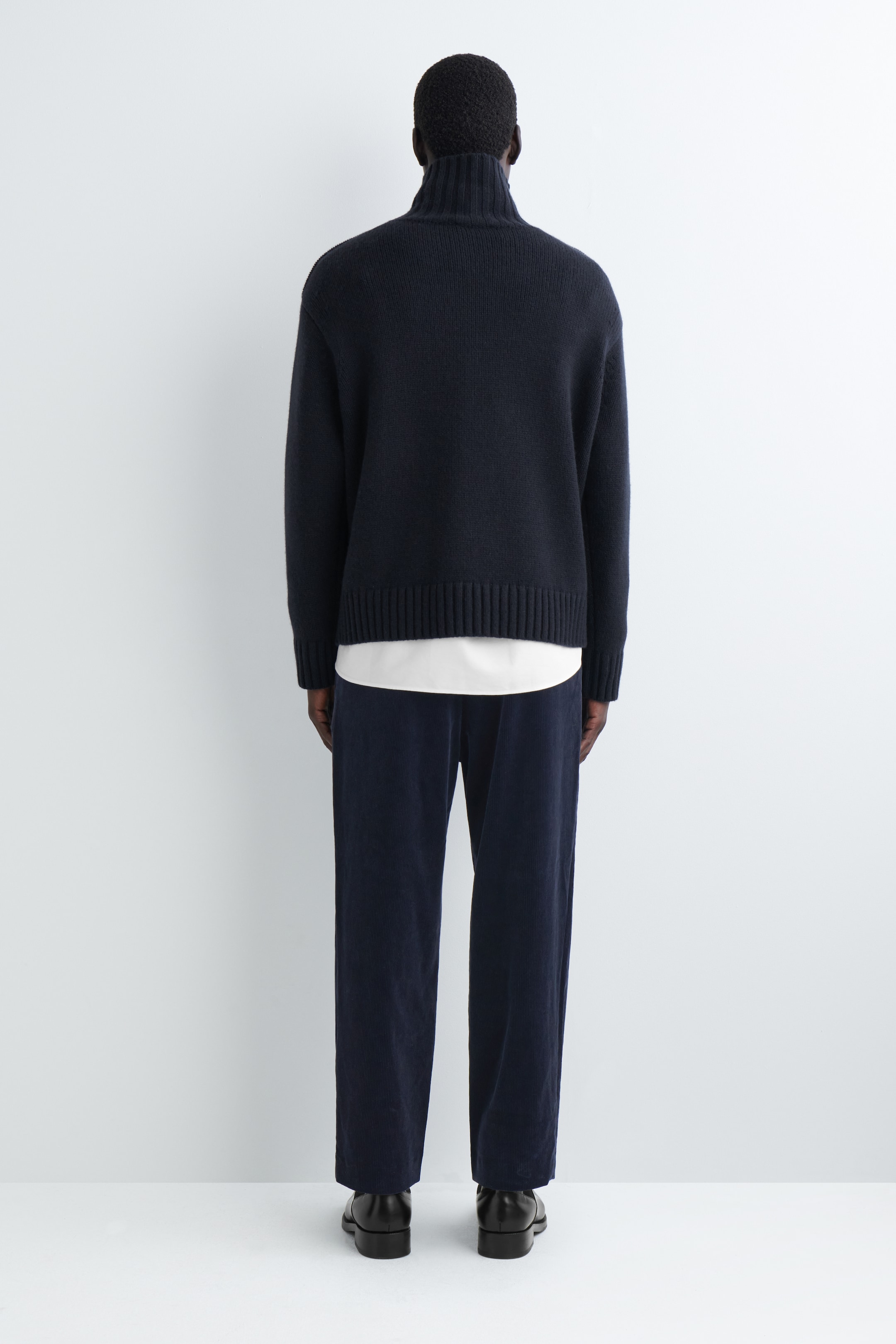 Größeres Bild ansehen: Ein dunkelblauer Strickpullover wird von hinten gezeigt, mit einem gerippten Stehkragen und einem breiten gerippten Saum, getragen über einem sichtbaren weißen Rundhals-T-Shirt. Passende dunkelblaue Cordhosen mit weitem Bein vervollständigen den Look.