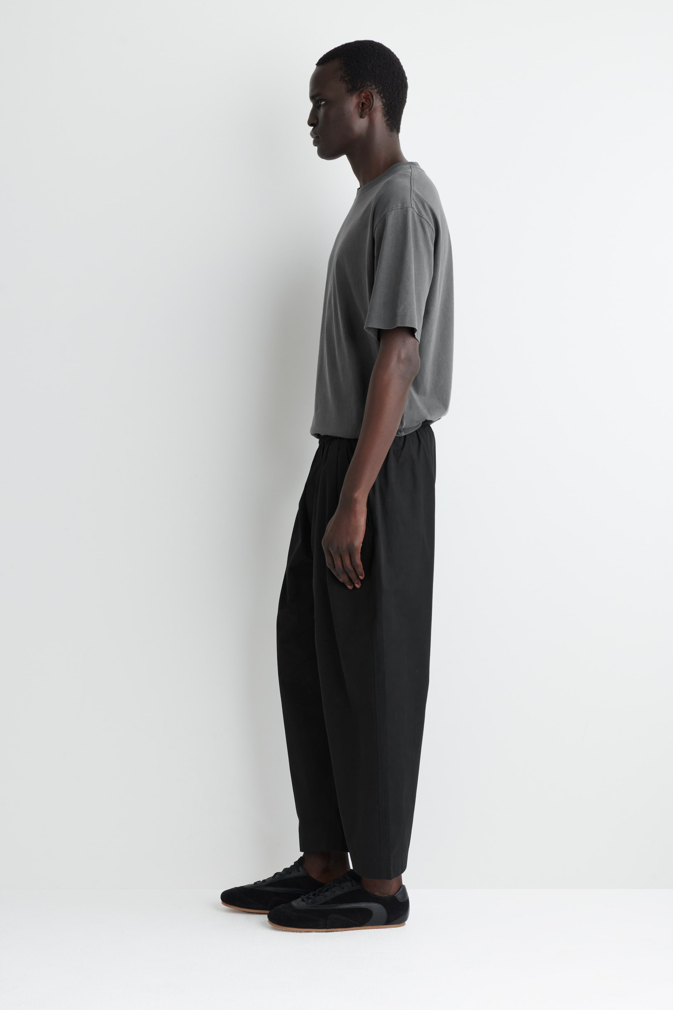 Agrandir l'image: Un t-shirt à col rond gris foncé, coupe ample, est porté avec son ourlet rentré dans un pantalon noir à jambes larges, doté d'une taille élastiquée et créant une silhouette froncée. Des baskets en daim noir avec des semelles en gomme complètent l'ensemble, le tout présenté de profil.