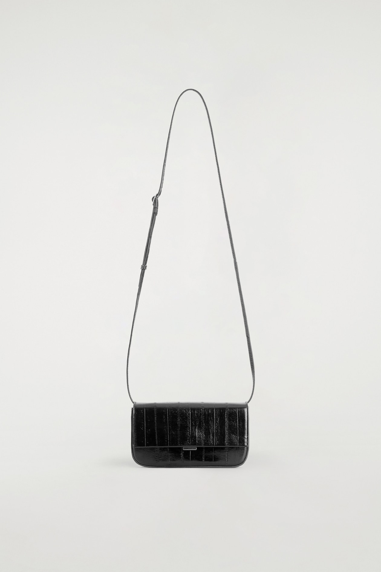 The Parquet Shoulder Bag in black eel-effect leather is shown from the front, showcasing its structured rectangular shape and glossy, vertically panelled texture. It features a front flap closure with a minimal metal clasp and is suspended by a long, thin, adjustable shoulder strap.