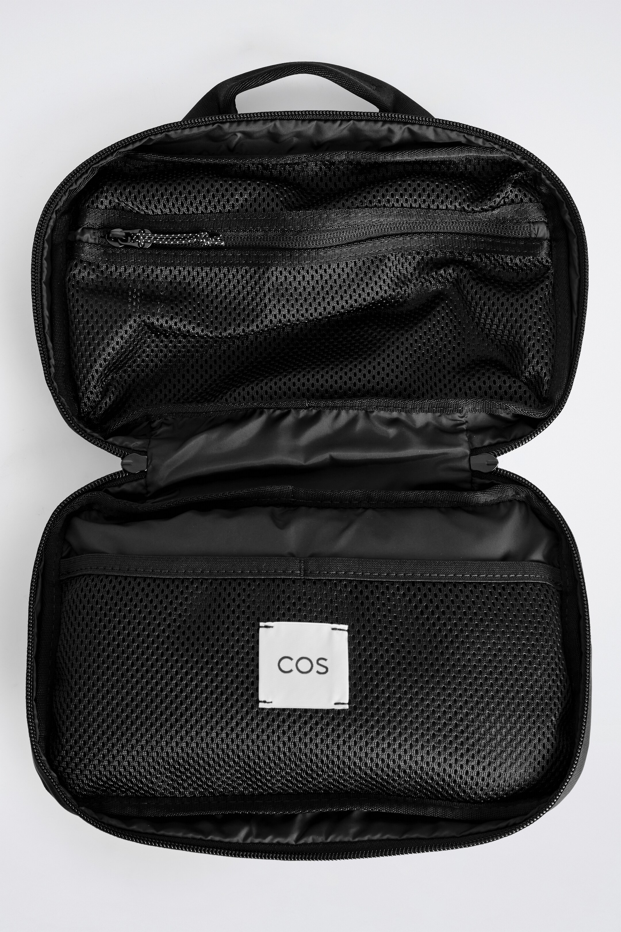 Agrandir l'image: Un sac de lavage noir est ouvert, vu de haut, mettant en valeur son organisation intérieure. La moitié supérieure comporte deux poches en maille noire, l'une avec une tirette de fermeture éclair détaillée réfléchissante, tandis que la moitié inférieure affiche une grande poche en maille noire avec une étiquette blanche 'COS'.
