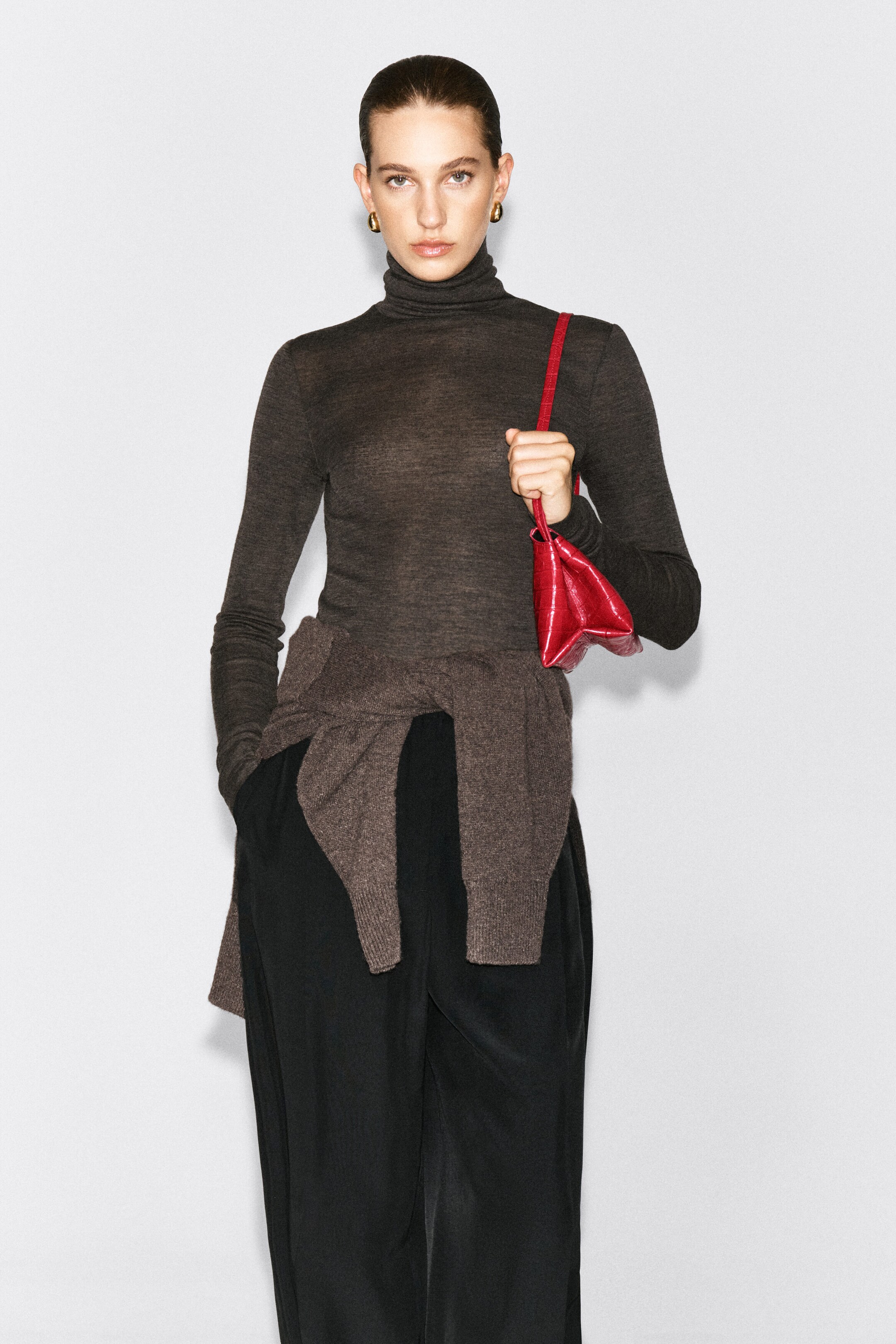 A merino wool high-neck top in dark brown is being worn by the model, featuring a fine-knit, semi-sheer texture and a slim fit. The top is styled with black trousers, a matching brown sweater tied around the waist, and a red shoulder bag worn on the shoulder. A merino wool high-neck top in dark brown is being worn by the model, featuring a fine-knit, semi-sheer texture and a slim fit. The top is styled with black trousers, a matching brown sweater tied around the waist, and a red shoulder bag worn on the shoulder.
